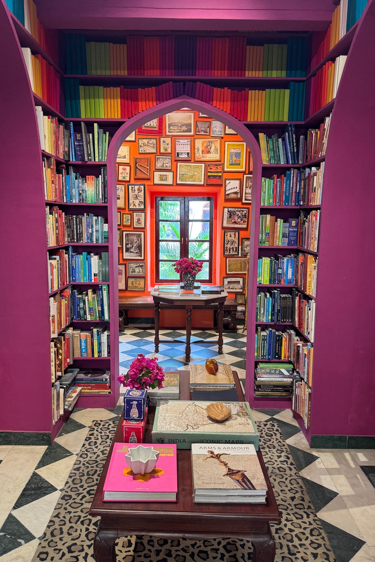 A cozy, highly decorated library interior with tall bookshelves lining magenta-purple walls, filled with colorful books. An arched doorway in the center leads to a small round table and window.