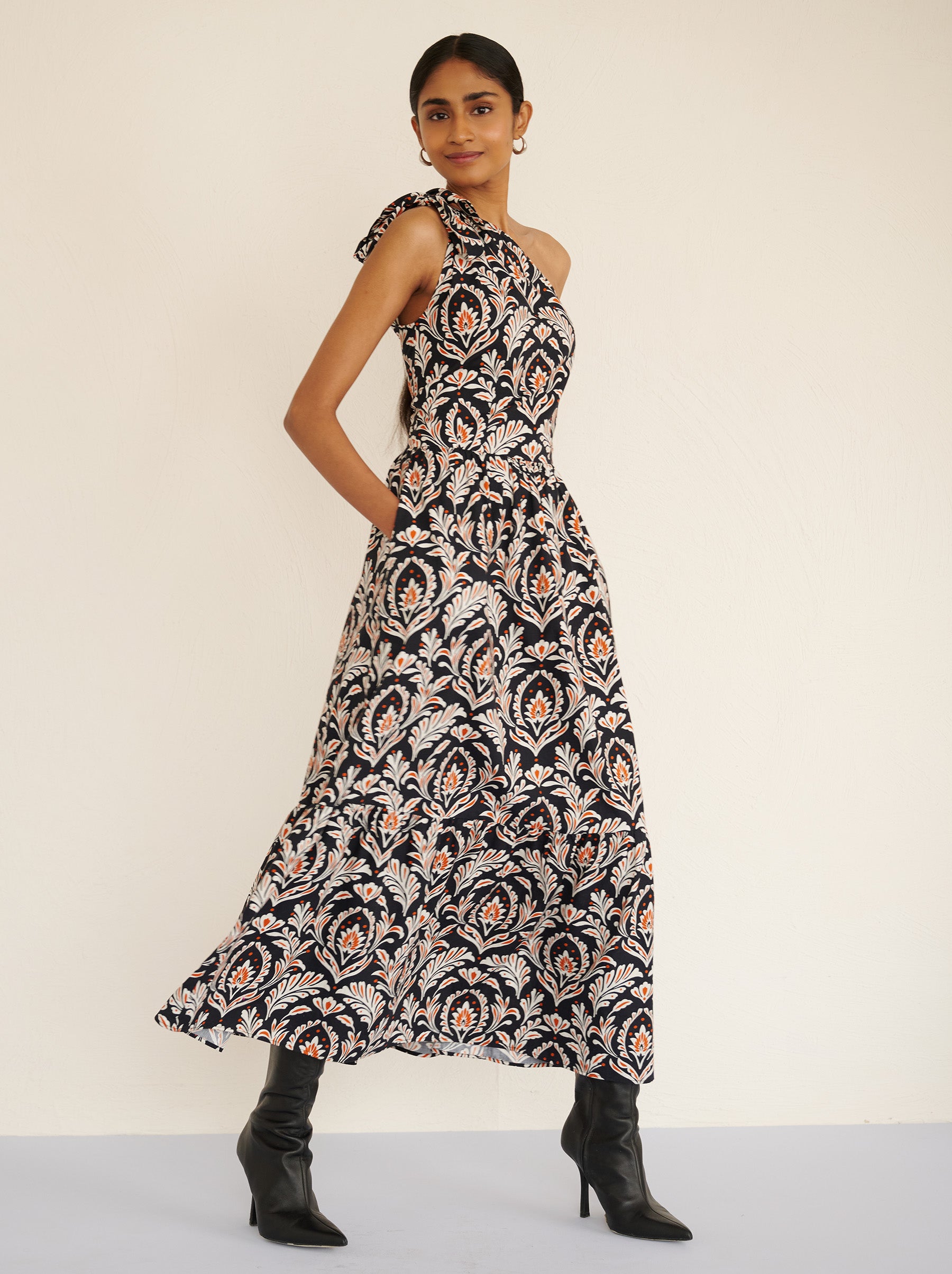 Side view of a model walking. She is wearing the Aliza full-length dress in crisp cotton poplin with the Akbar print. It is featuring an adjustable tie at the shoulder and side-seam pockets.