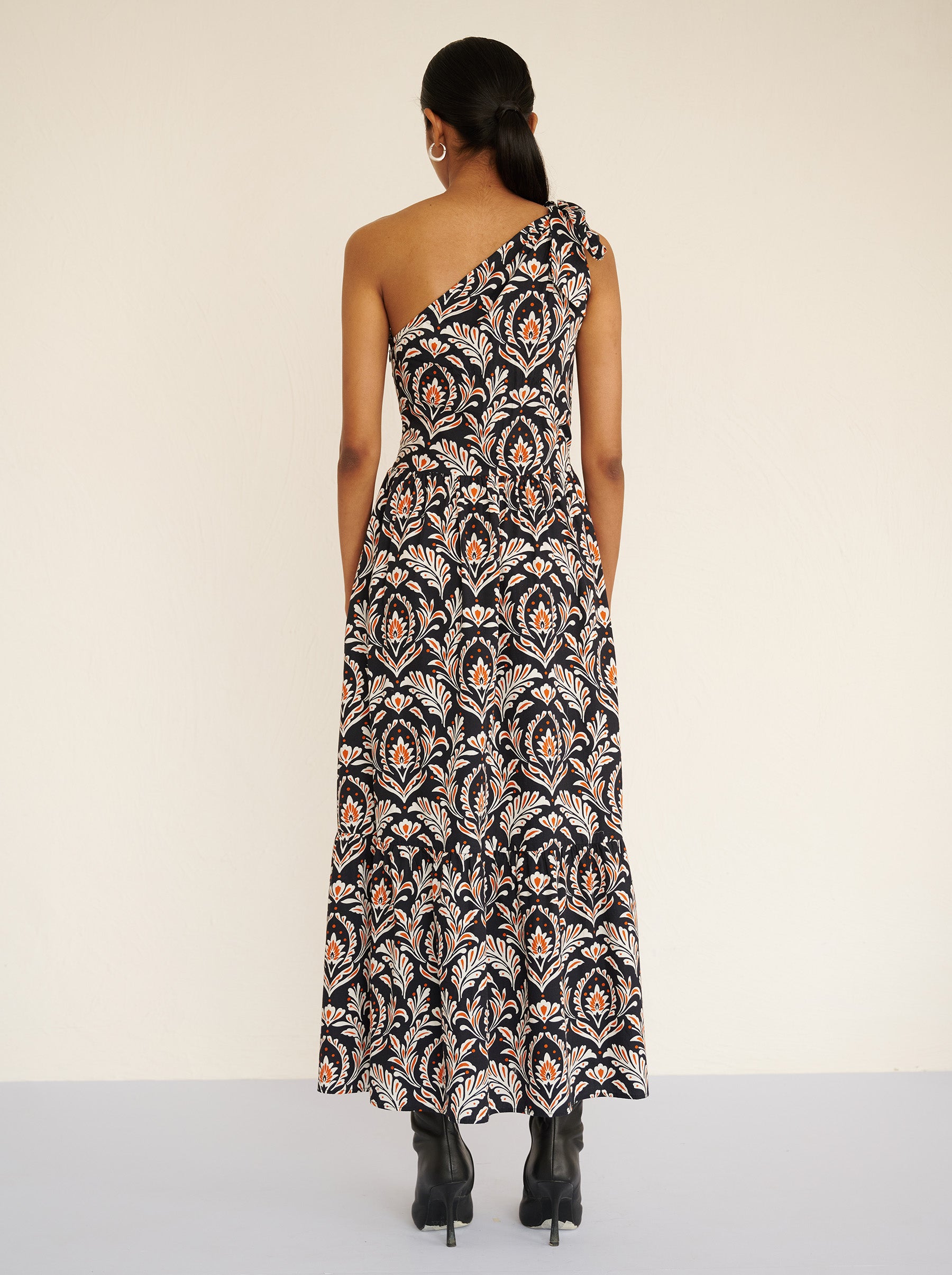 Back view of a model standing. She is wearing the Aliza full-length dress in crisp cotton poplin with the Akbar print. It is featuring an adjustable tie at the shoulder and side-seam pockets.