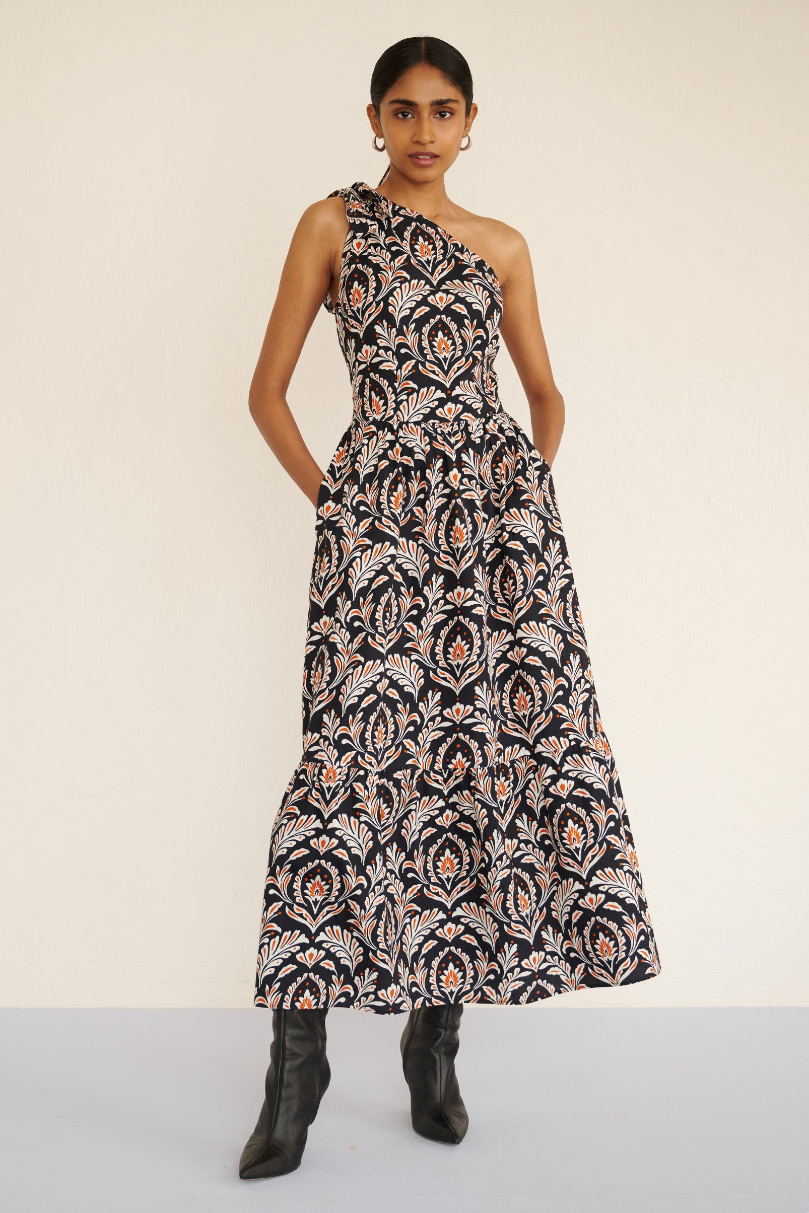 Front view of a model standing with hands in pockets. She is wearing the Aliza full-length dress in crisp cotton poplin with the Akbar print. It is featuring an adjustable tie at the shoulder and side-seam pockets.