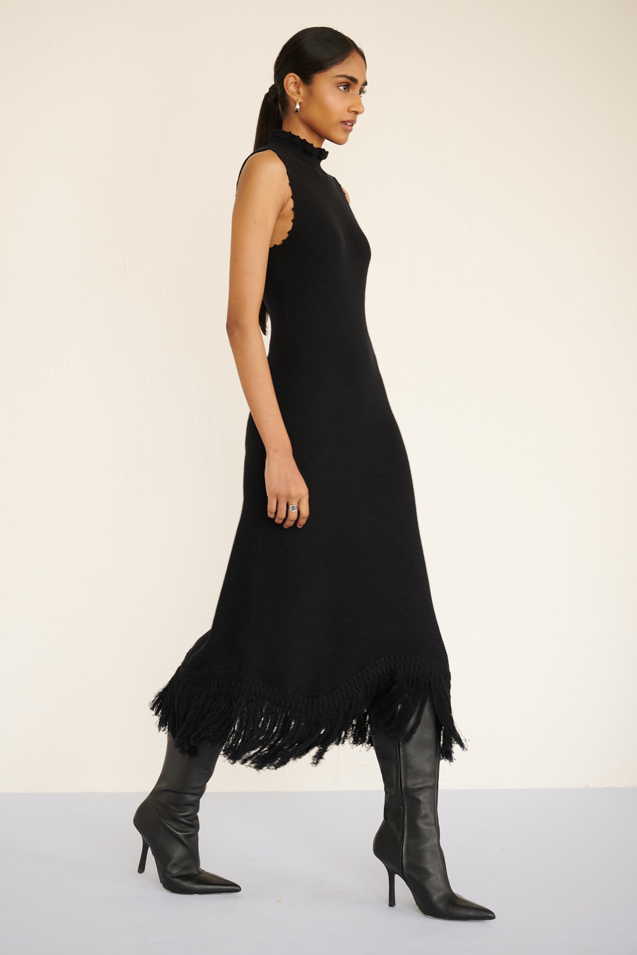 Side view of woman wearing a sleeveless cotton midi dress with a high neckline, hand-crocheted trim, and fringed asymmetrical hem.