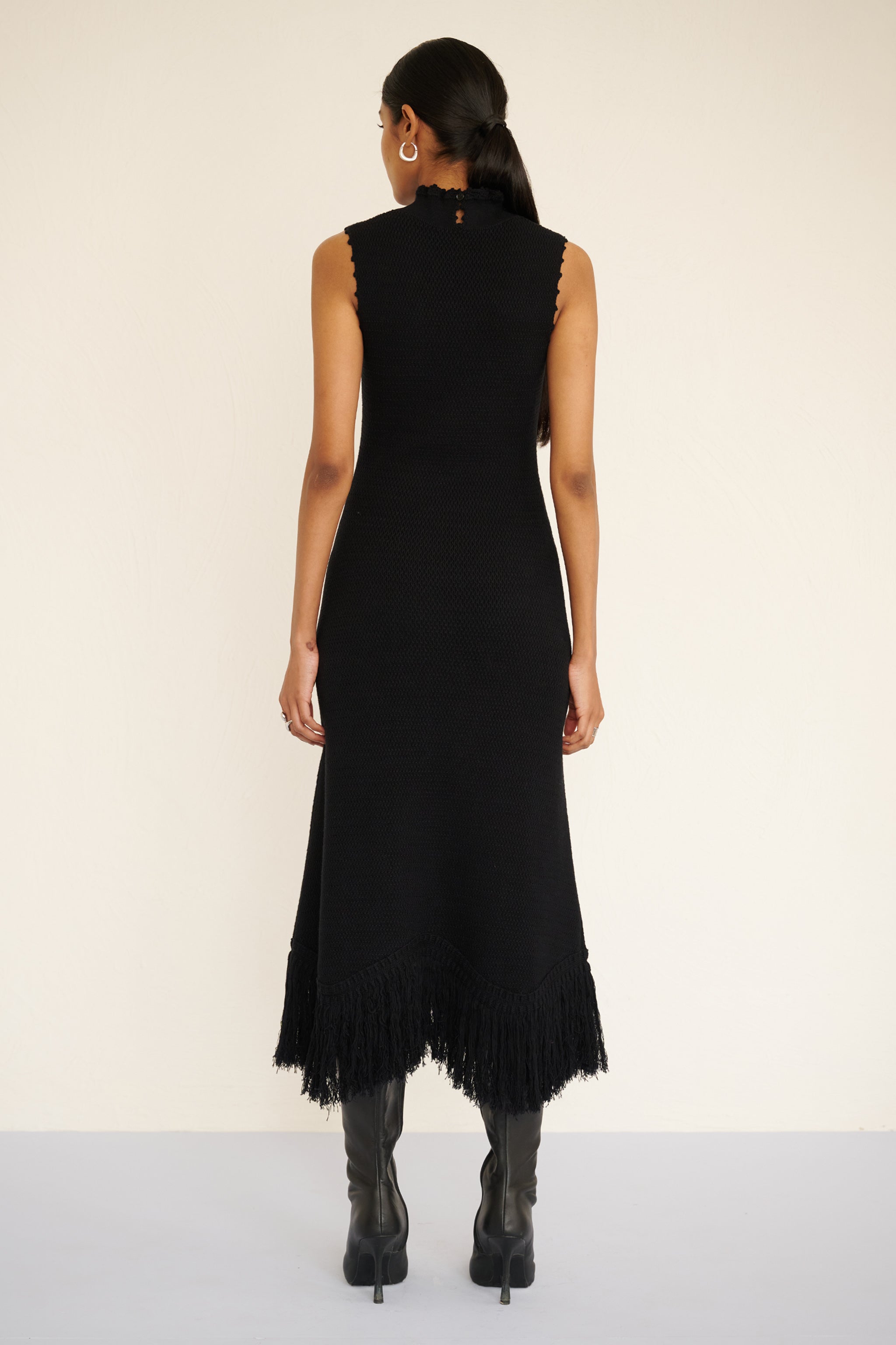 Back view of woman wearing a sleeveless cotton midi dress with a high neckline, hand-crocheted trim, and fringed asymmetrical hem.
