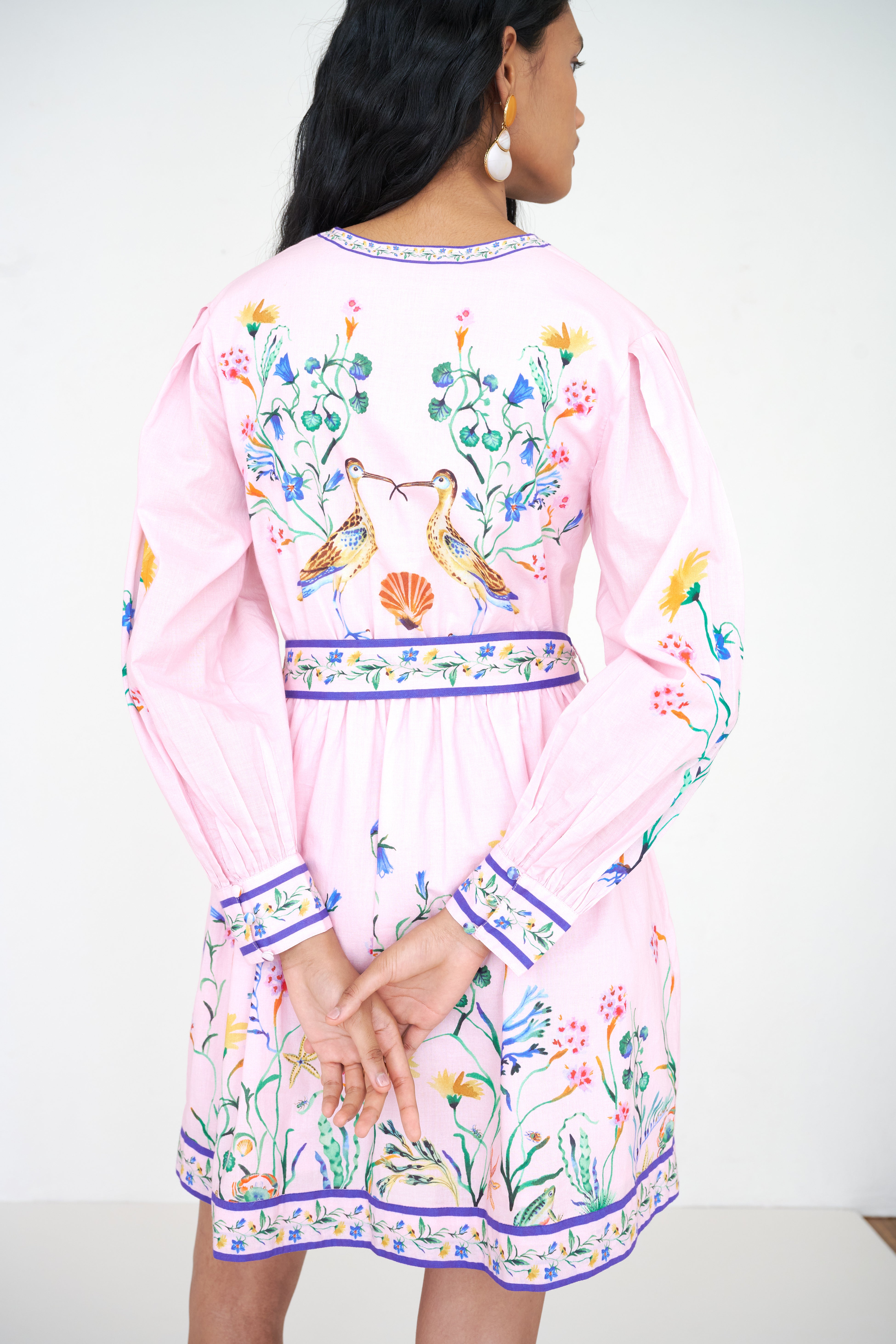 Back view close up of a woman standing wearing the Anne dress in Tidepool print. It features long sleeves, elasticated waist, fabric-covered buttons with loops, fabric belt, side-seam pockets, and a short hemline.