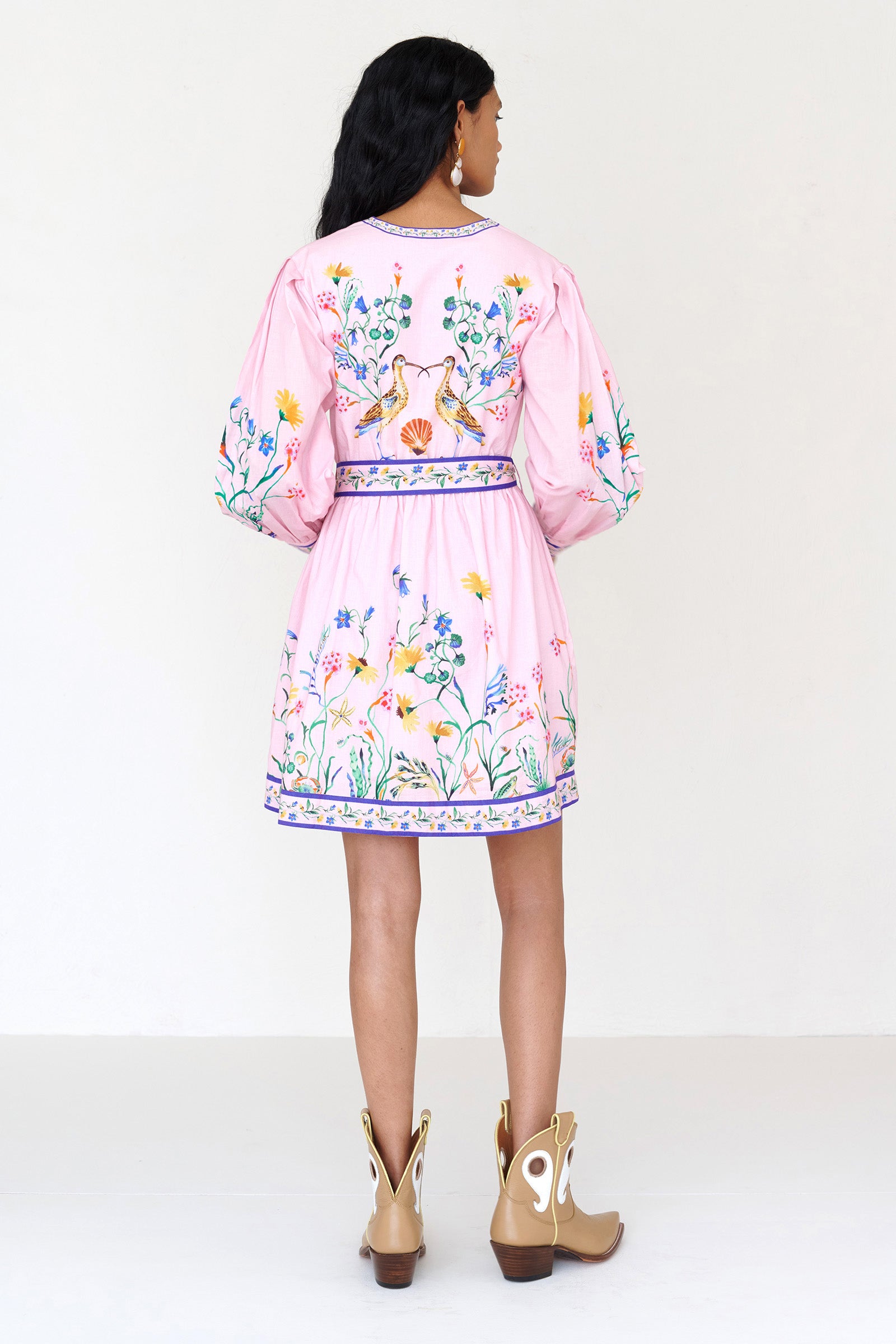 Back view of a woman standing wearing the Anne dress in Tidepool print. It features long sleeves, elasticated waist, fabric-covered buttons with loops, fabric belt, side-seam pockets, and a short hemline.