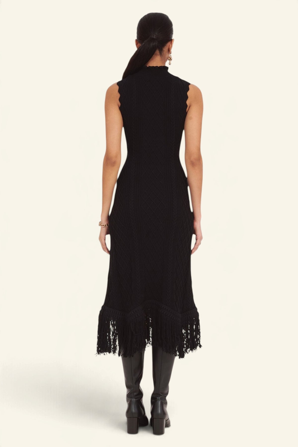 Back view of model standing wearing the Amelia Fringe Dress, featuring a body-skimming cable-knit silhouette, high neckline with hand-crocheted trim, and an uneven hem finished with fringe.