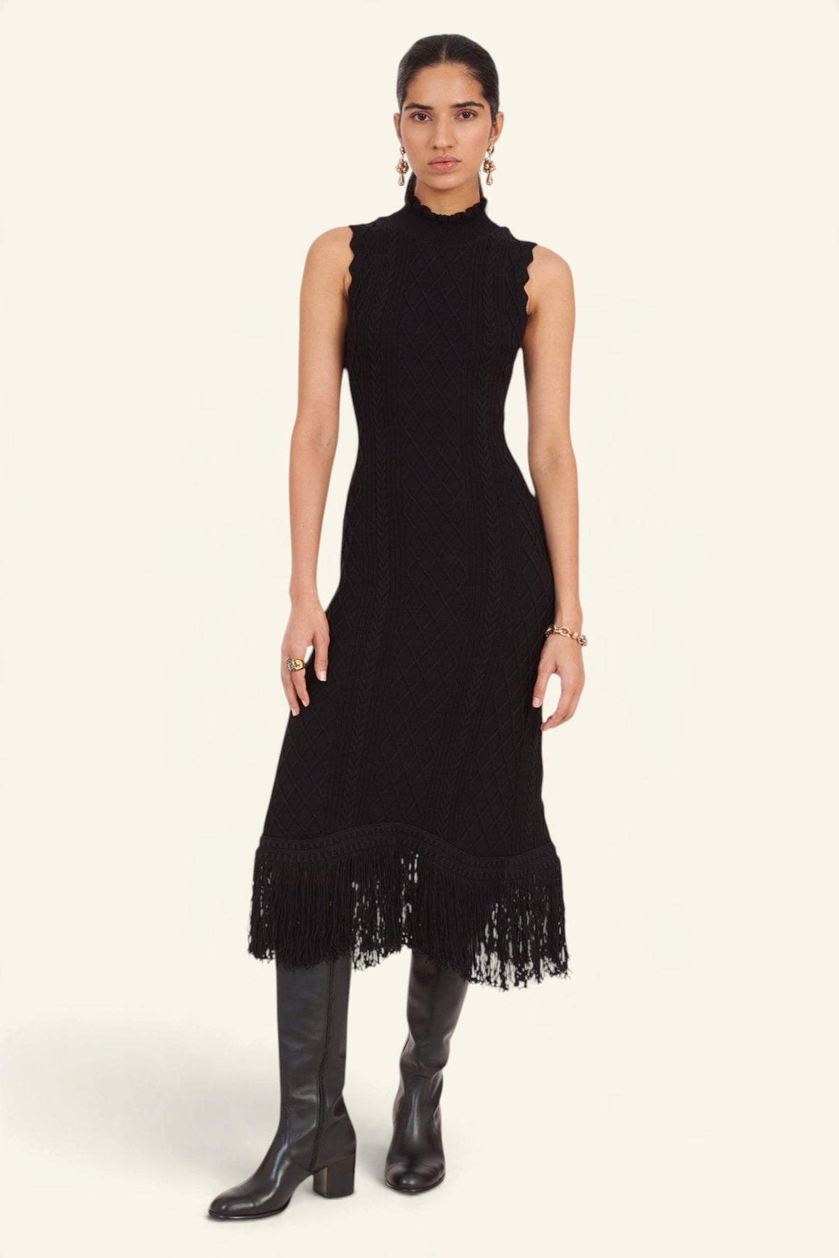 Front view of model standing wearing the Amelia Fringe Dress, featuring a body-skimming cable-knit silhouette, high neckline with hand-crocheted trim, and an uneven hem finished with fringe.