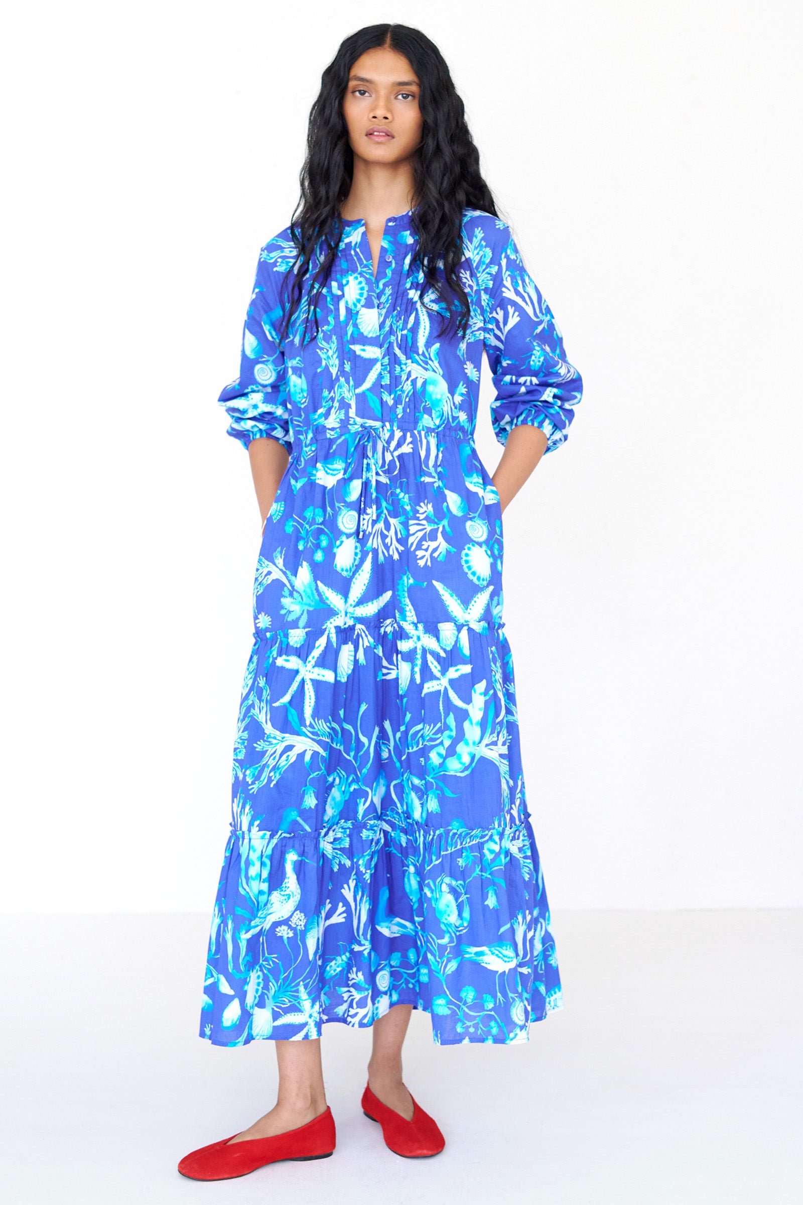 Front view of a woman standing wearing the Bazaar dress in Tidepool print. It features long sleeves, tiered ankle-length skirt, drawstring belt and side-seam pockets.