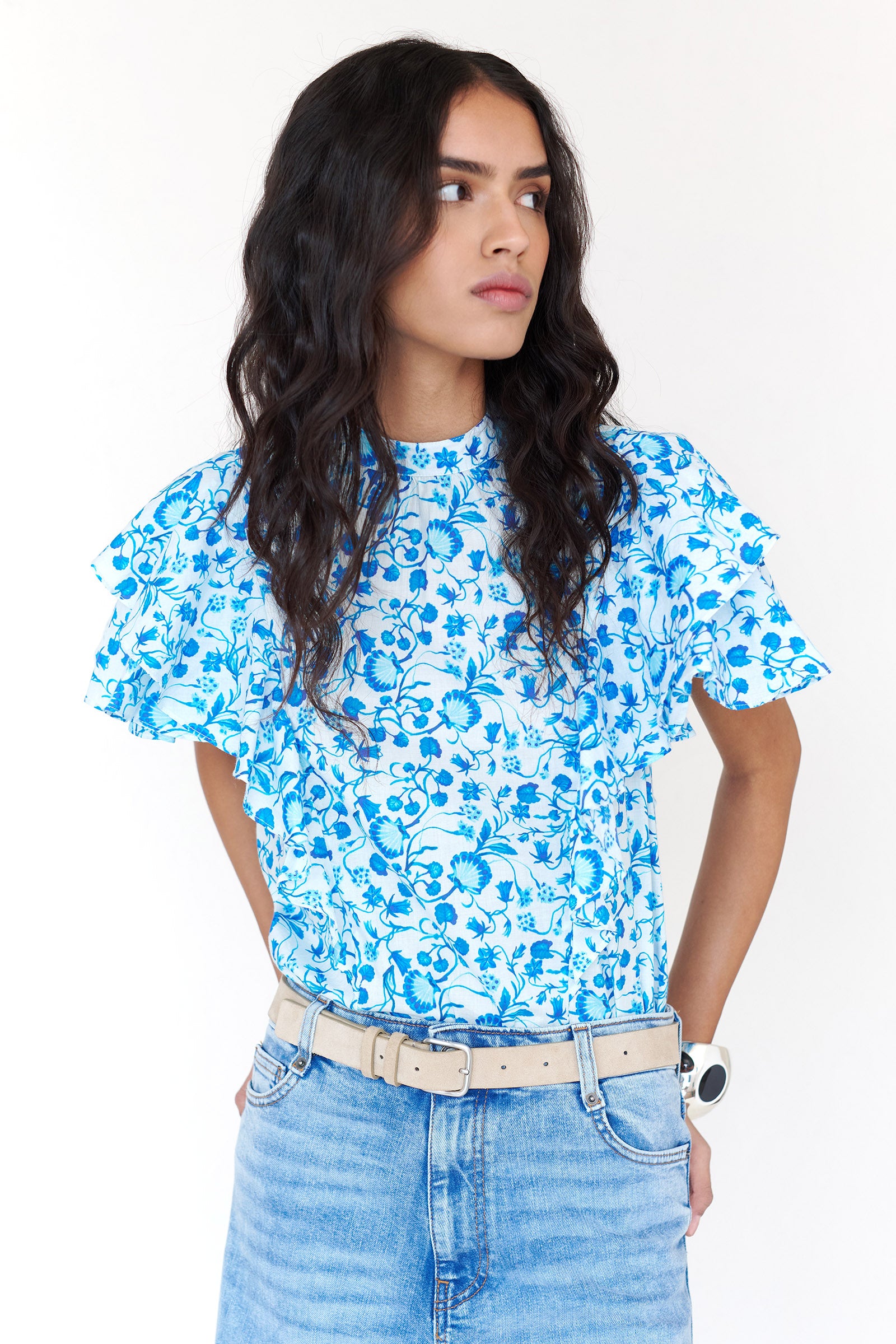 Front view of a woman standing. She is wearing the Bebe top in Clam Garland print. It features a soft shoulder drape, tie neckline, and layered sleeves. Crafted in a timeless floral inspired by the Scottish Isles, styled with denim.