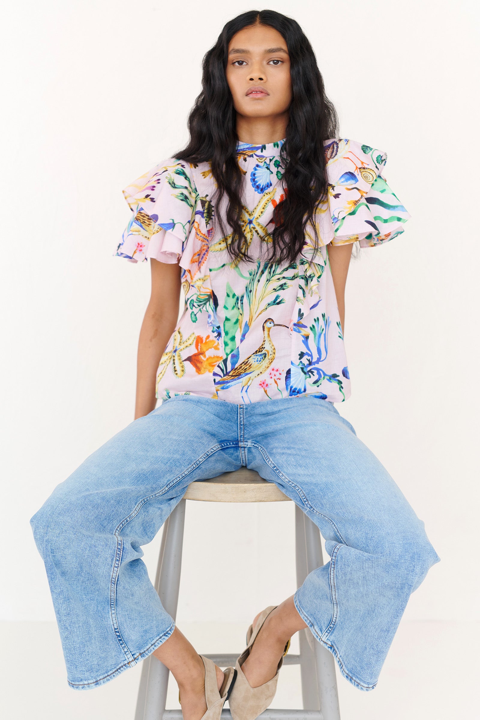 Front view of a woman sitting on a chair. She is wearing the Bebe top in Tidepool print, featuring a soft shoulder drape, tie neckline, and full layered sleeves. Ocean-inspired print, styled with denim.