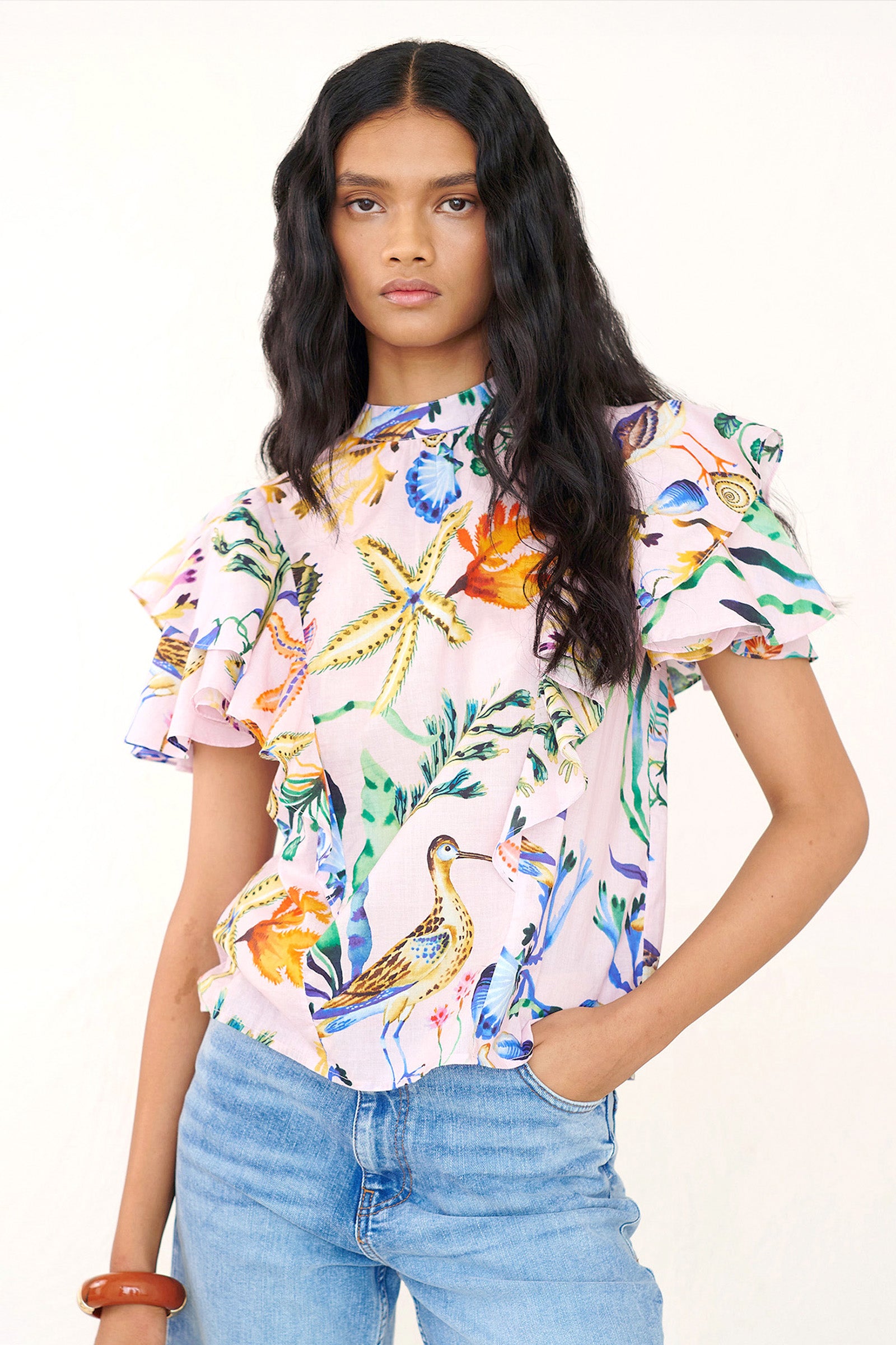 Front view of a woman standing wearing the Bebe top in Tidepool print, featuring a soft shoulder drape, tie neckline, and full layered sleeves. Ocean-inspired print, styled with denim.