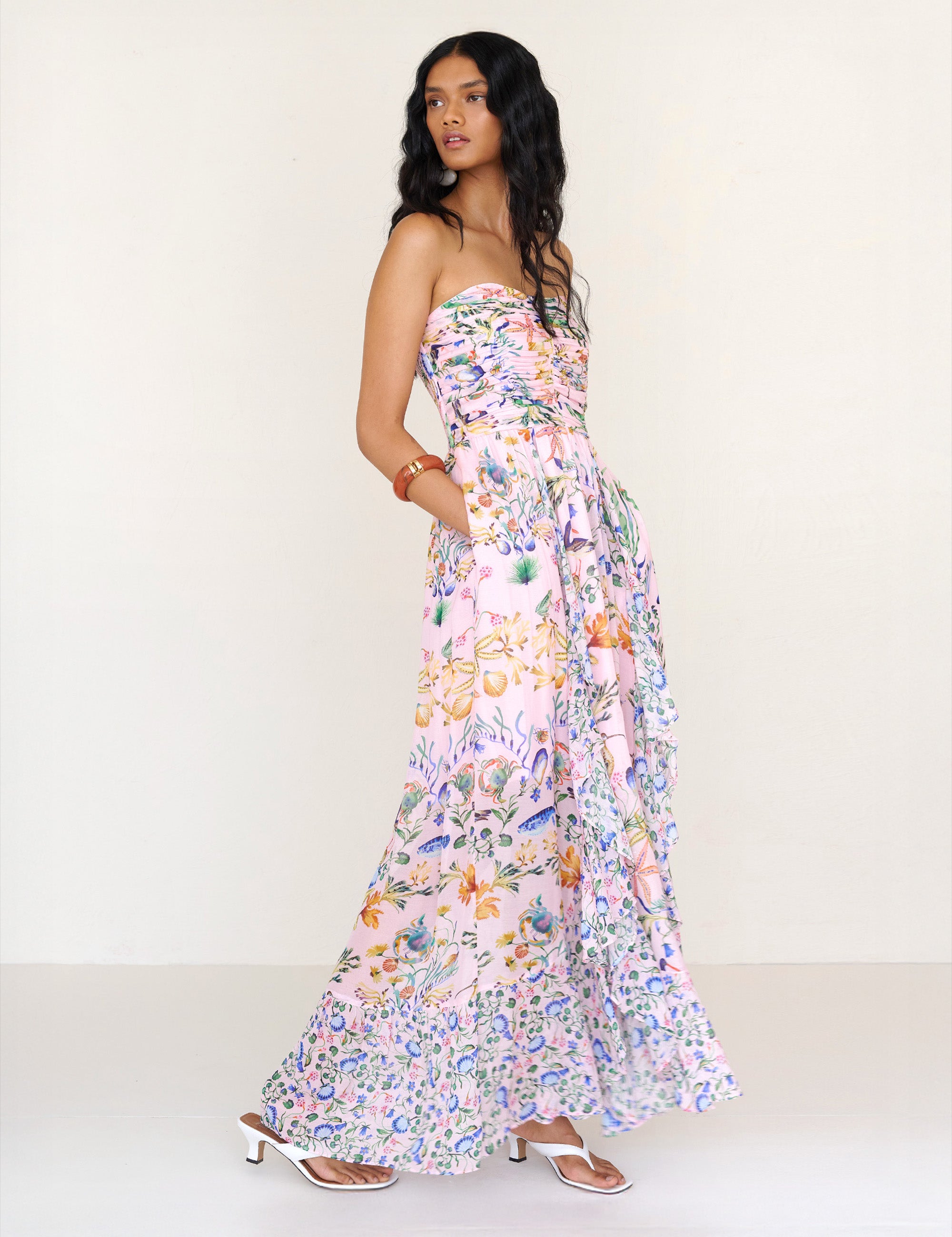 Side view of a woman standing with hands in pocket. She is wearing the Bella full-length dress in Tidepool print. It features a cascading overlapping skirt with frill detail in contrast conch print, ruched bodice, smocked back, and side-seam pockets.