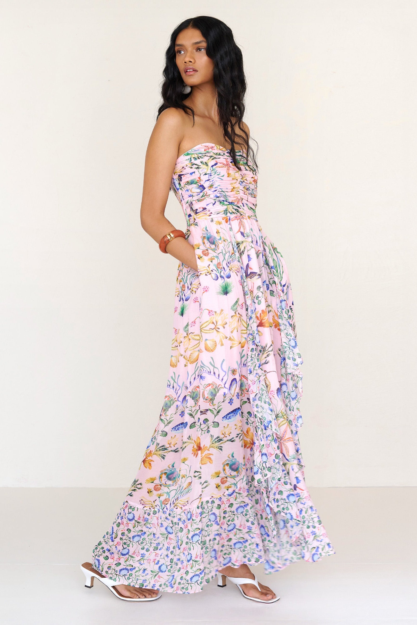 Side view of a woman standing with hands in pocket. She is wearing the Bella full-length dress in Tidepool print. It features a cascading overlapping skirt with frill detail in contrast conch print, ruched bodice, smocked back, and side-seam pockets.