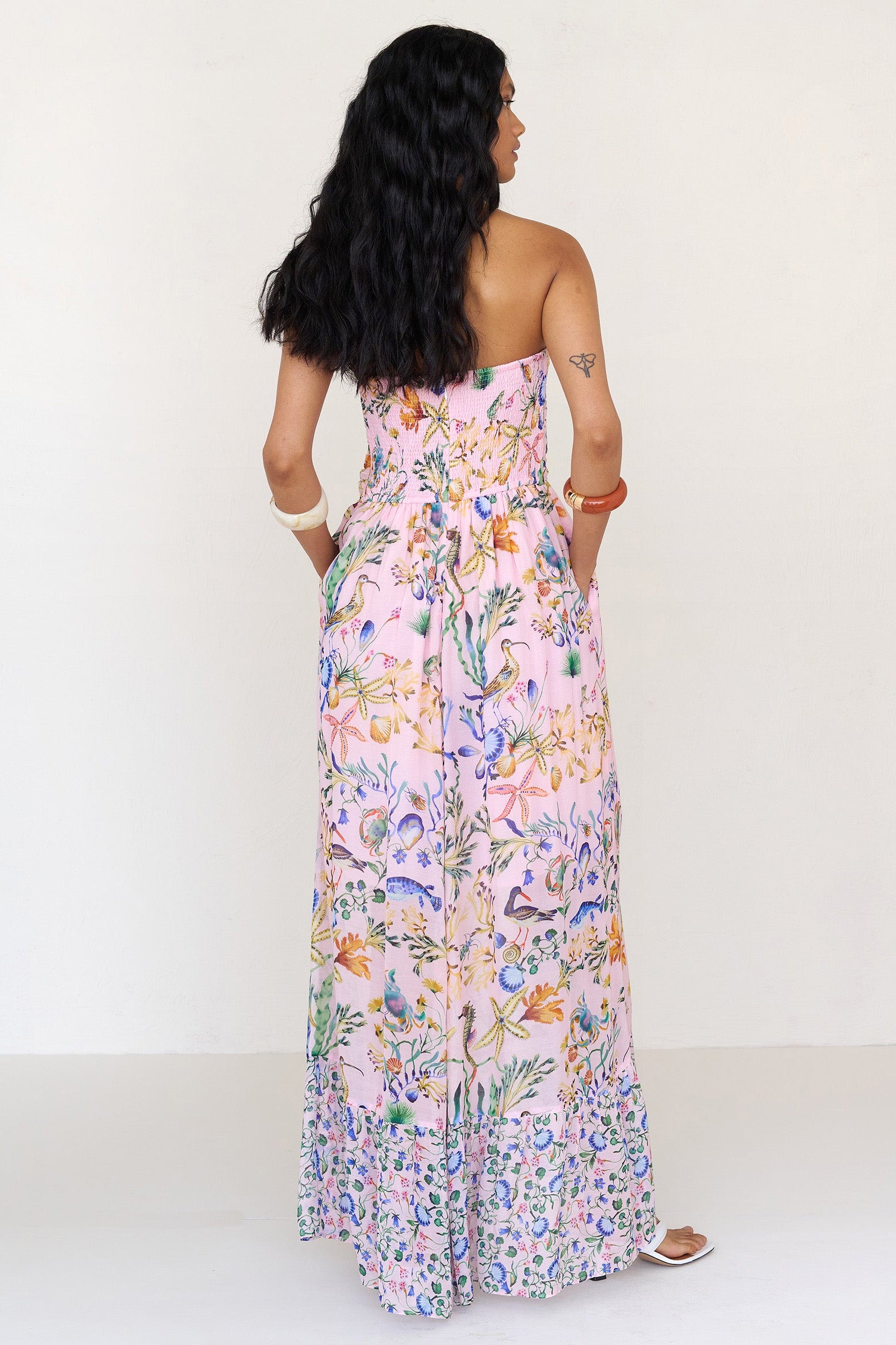 Back view of a woman standing wearing the Bella dress in Tidepool print, featuring a full-length cascading skirt with contrast frill, ruched bodice, smocked back, side-seam pockets, and drapey viscose voile.