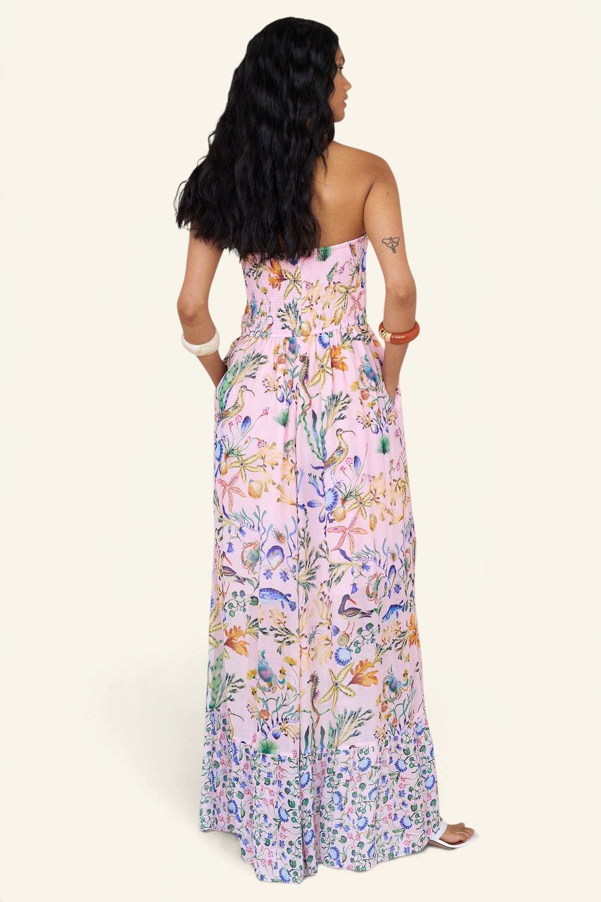 Back view of a woman standing wearing the Bella dress in Tidepool print, featuring a full-length cascading skirt with contrast frill, ruched bodice, smocked back, side-seam pockets, and drapey viscose voile.