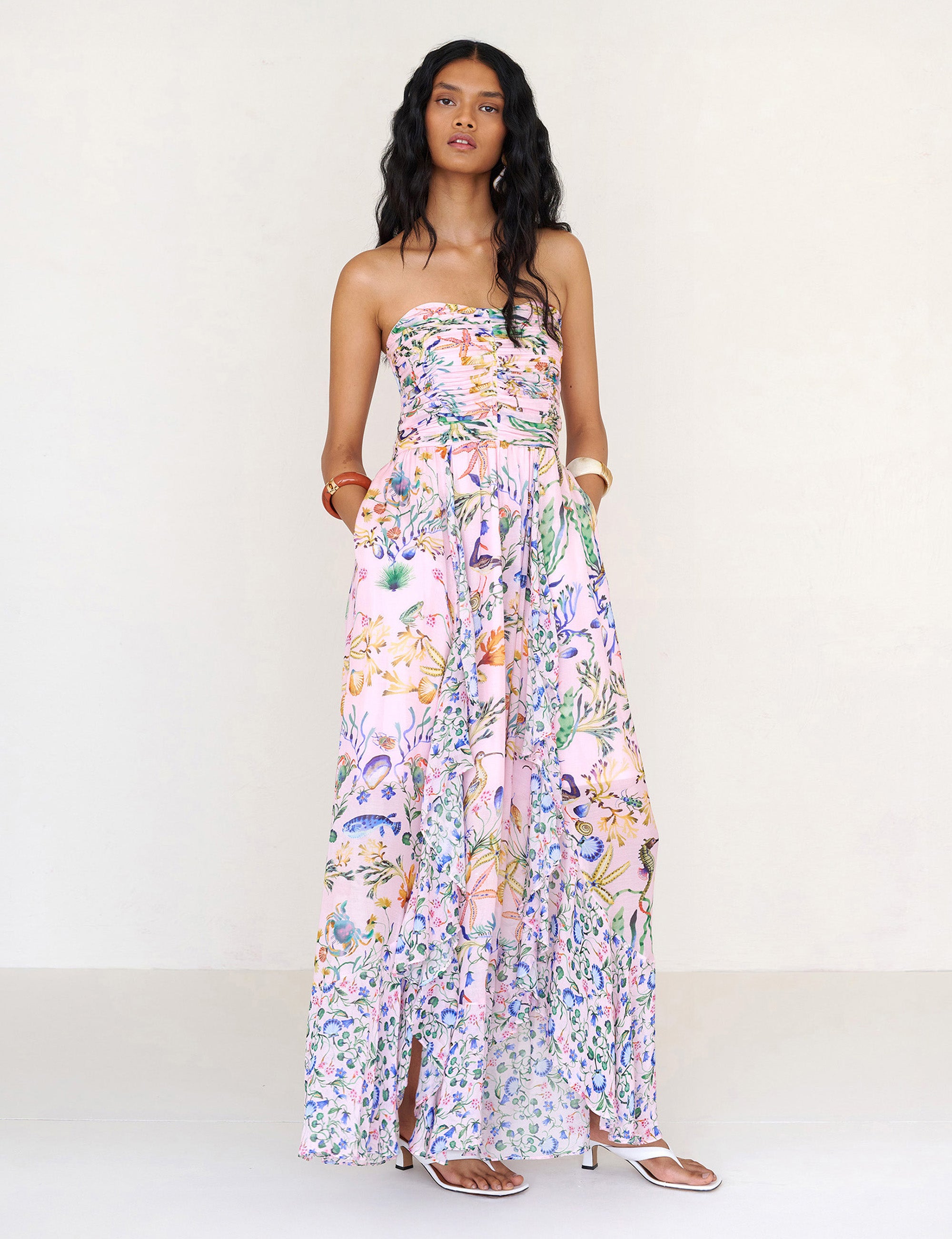 Front view of a woman standing with hands in pocket. She is wearing the Bella full-length dress in Tidepool print. It features a cascading overlapping skirt with frill detail in contrast conch print, ruched bodice, smocked back, and side-seam pockets.