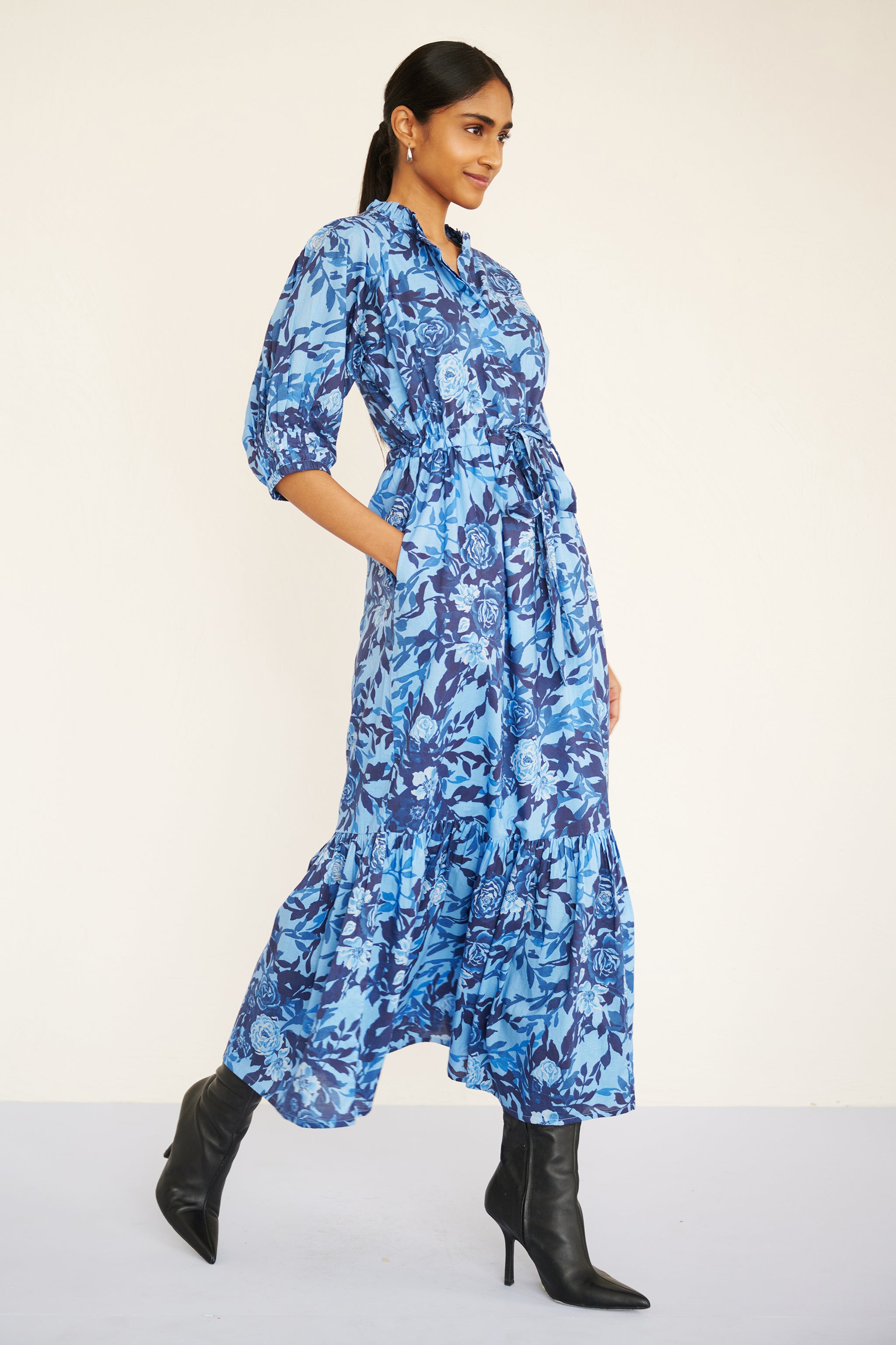 Side view of woman wearing a floral midi dress with raglan sleeves, gathered skirt, tie belt at the waist, and a soft watercolor rose print in tones of blue.