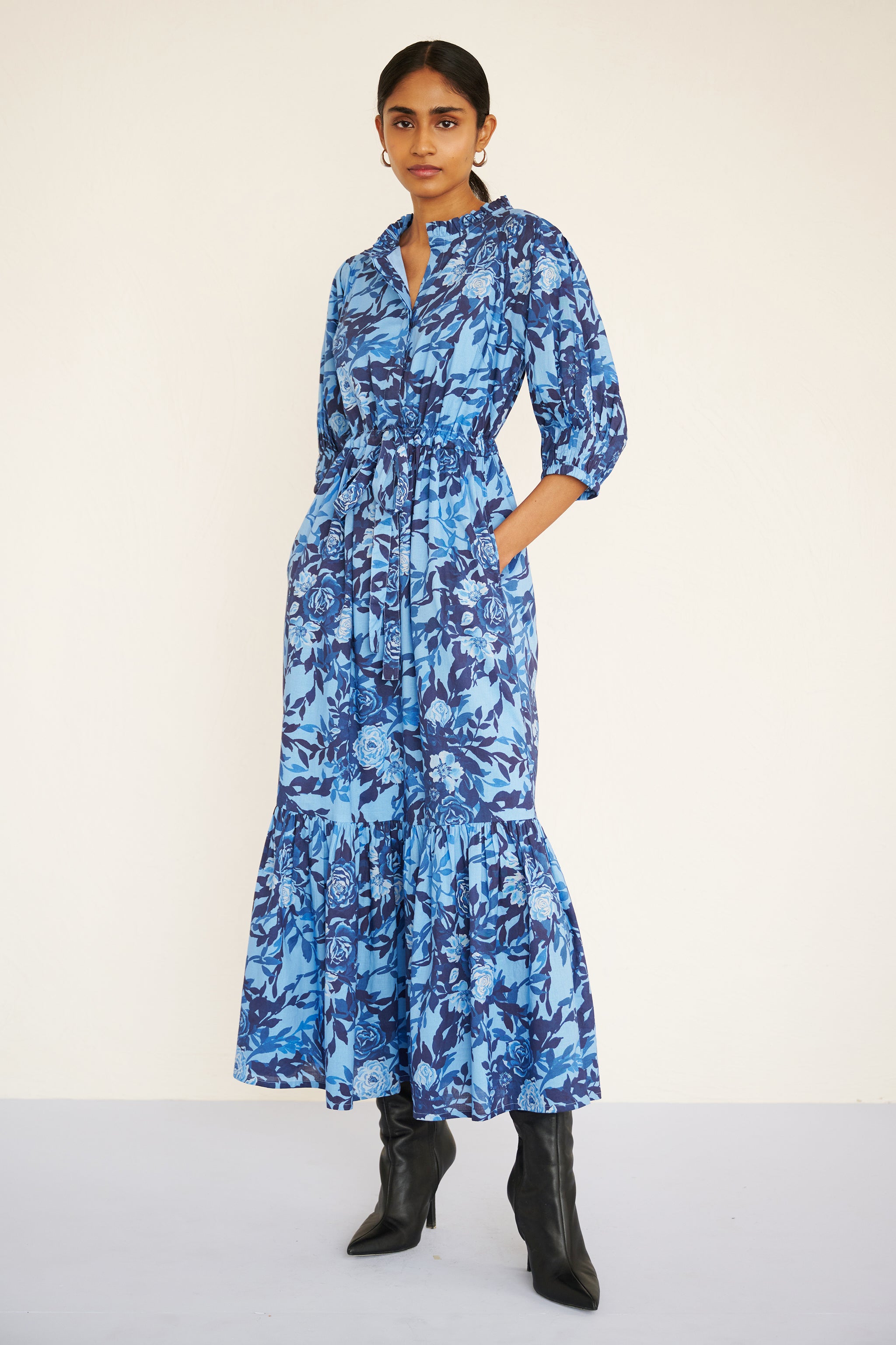 Woman wearing a floral midi dress with raglan sleeves, gathered skirt, tie belt at the waist, and a soft watercolor rose print in tones of blue.