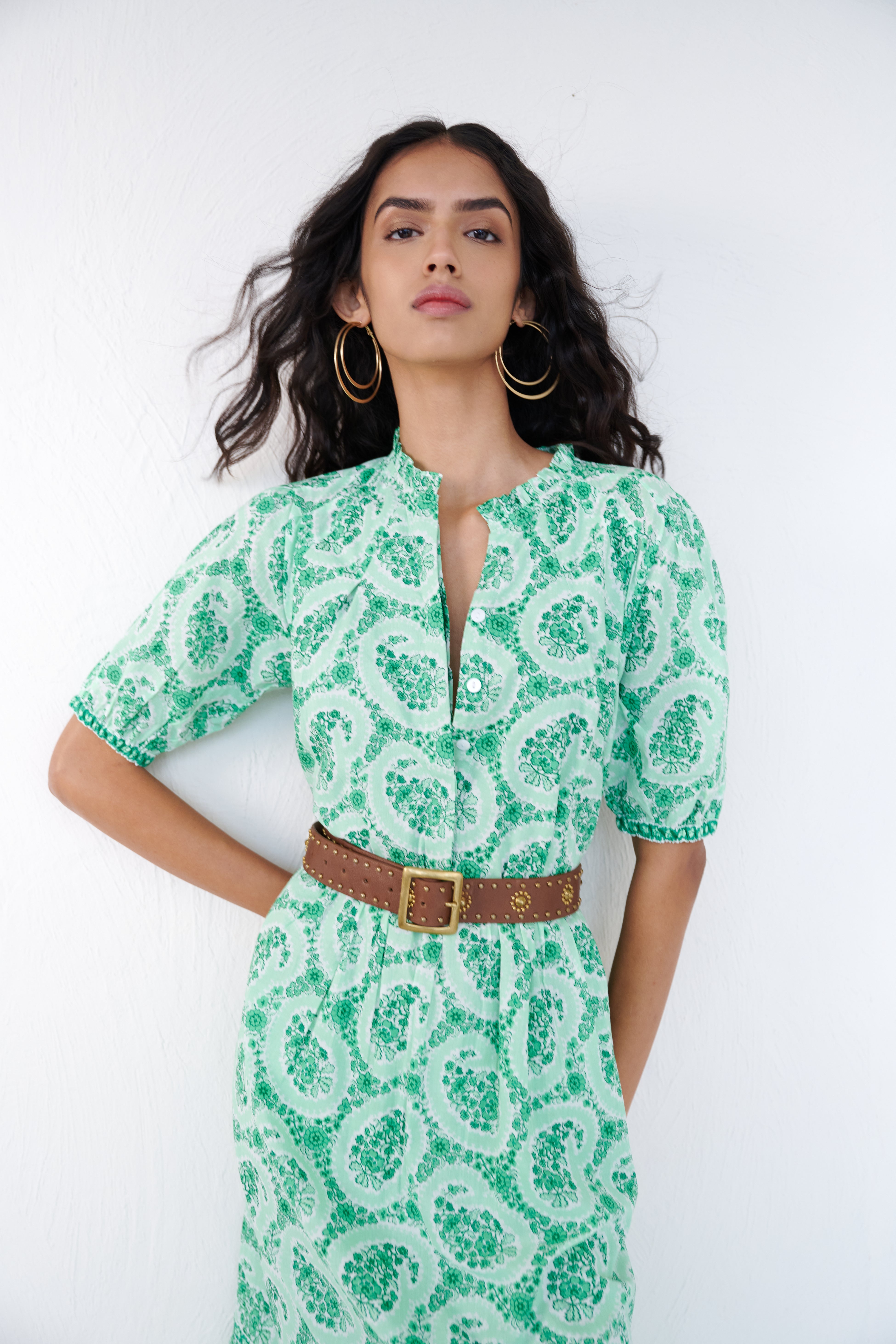 Front view close up of a woman standing wearing the Betty dress in Paisley Copse print. It features gathered skirt, raglan sleeves, contrast trim and side-seam pockets in breezy cotton voile. Styled with brown belt and gold accessories.