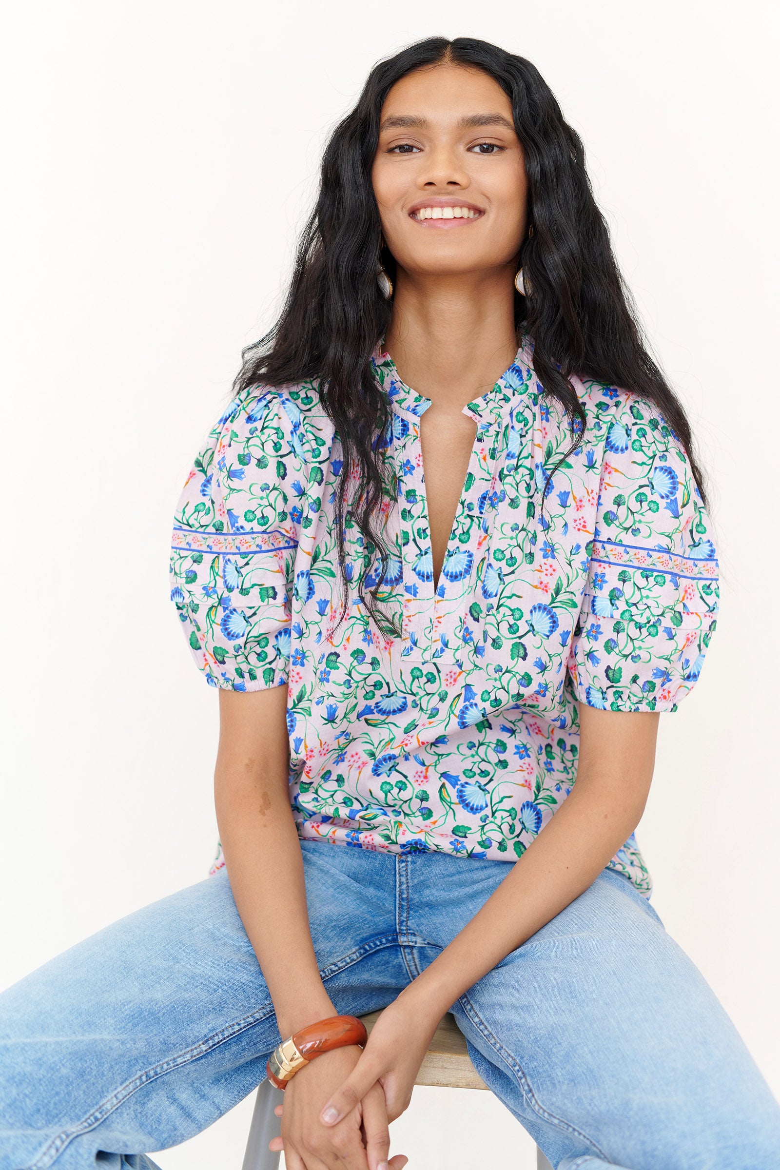 Front view of a woman sitting on a chair. She is wearing the Bronte top in Clam Garland print, featuring a front opening, delicate neckline detailing, contrast trim, and softly pleated sleeves styled with denim.