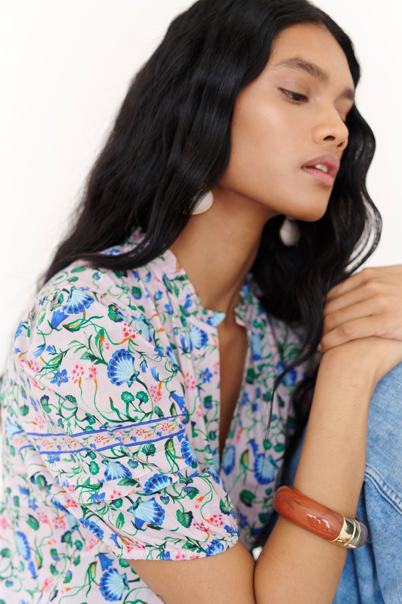 Side view close up of a woman sitting on a chair. She is wearing the Bronte top in Clam Garland print, featuring softly pleated sleeves styled with denim.