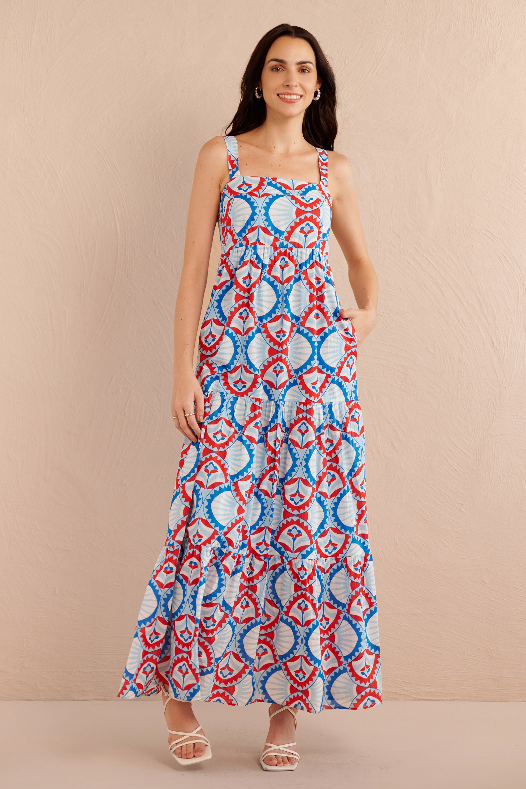 Front view of woman wearing a sleeveless tiered maxi dress with bold red, blue, and white geometric print, styled with white strappy heels.
