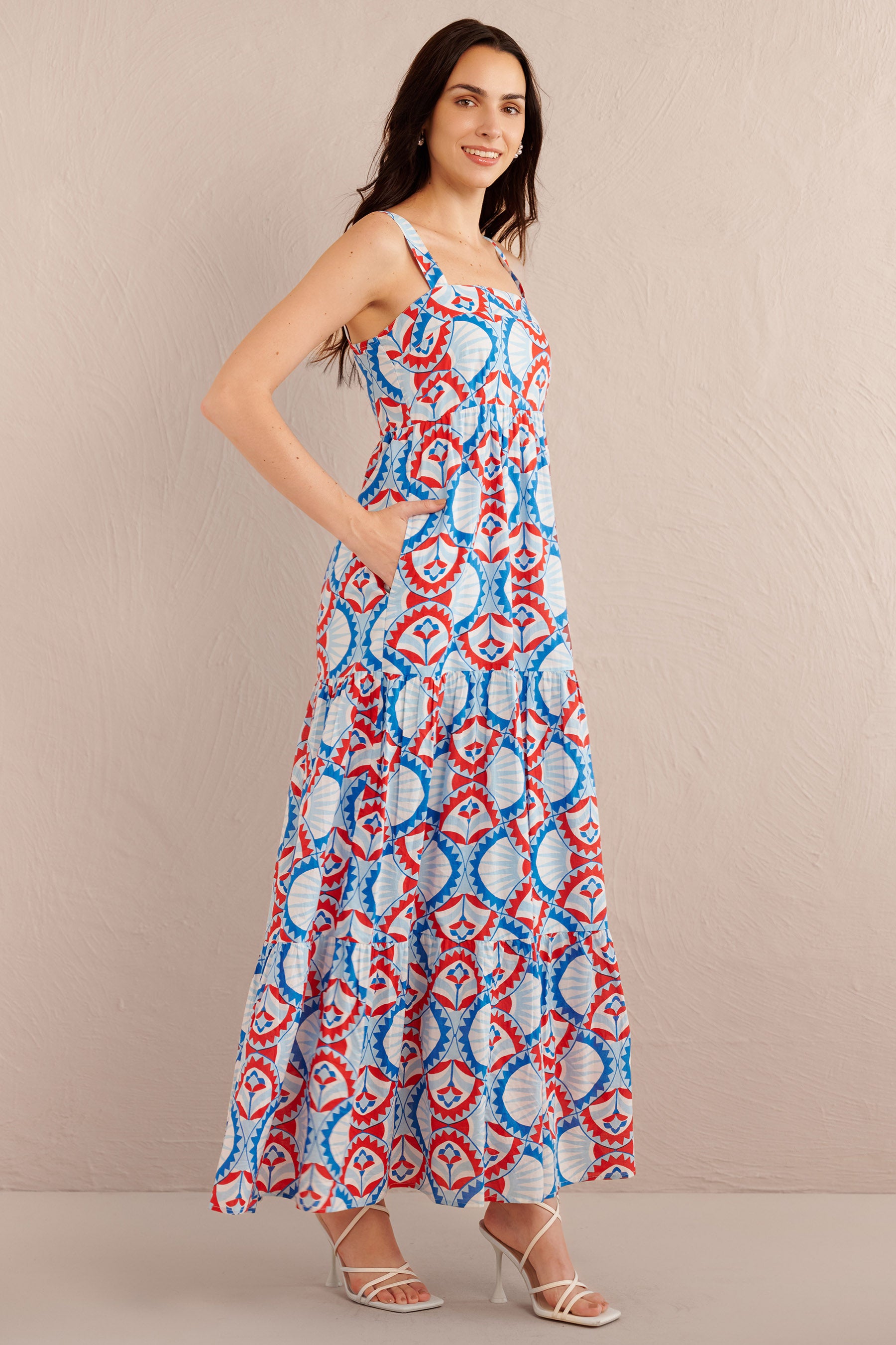 Side view of woman wearing a sleeveless tiered maxi dress with bold red, blue, and white geometric print, styled with white strappy heels.