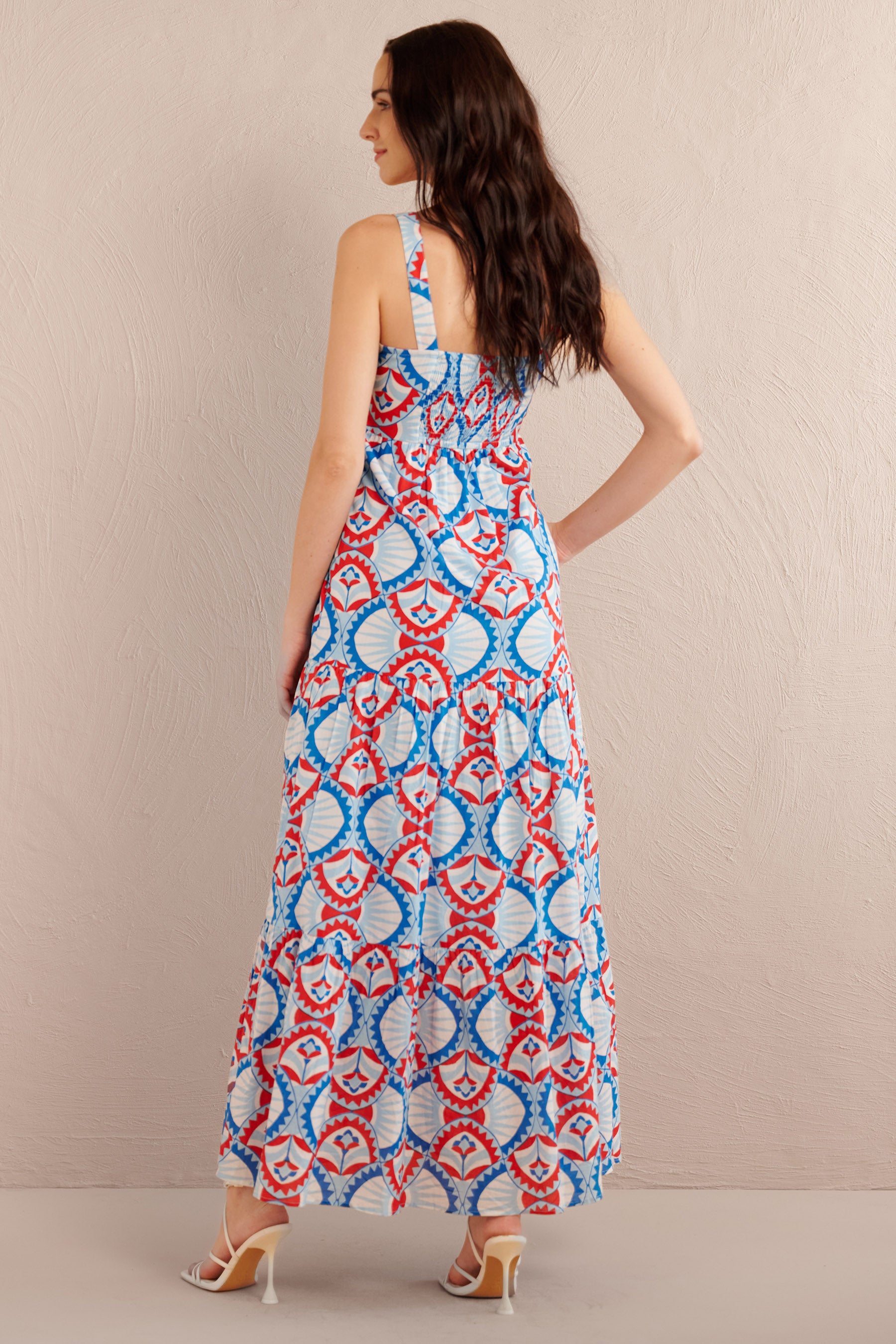 Back view of woman wearing a sleeveless tiered maxi dress with bold red, blue, and white geometric print, styled with white strappy heels.