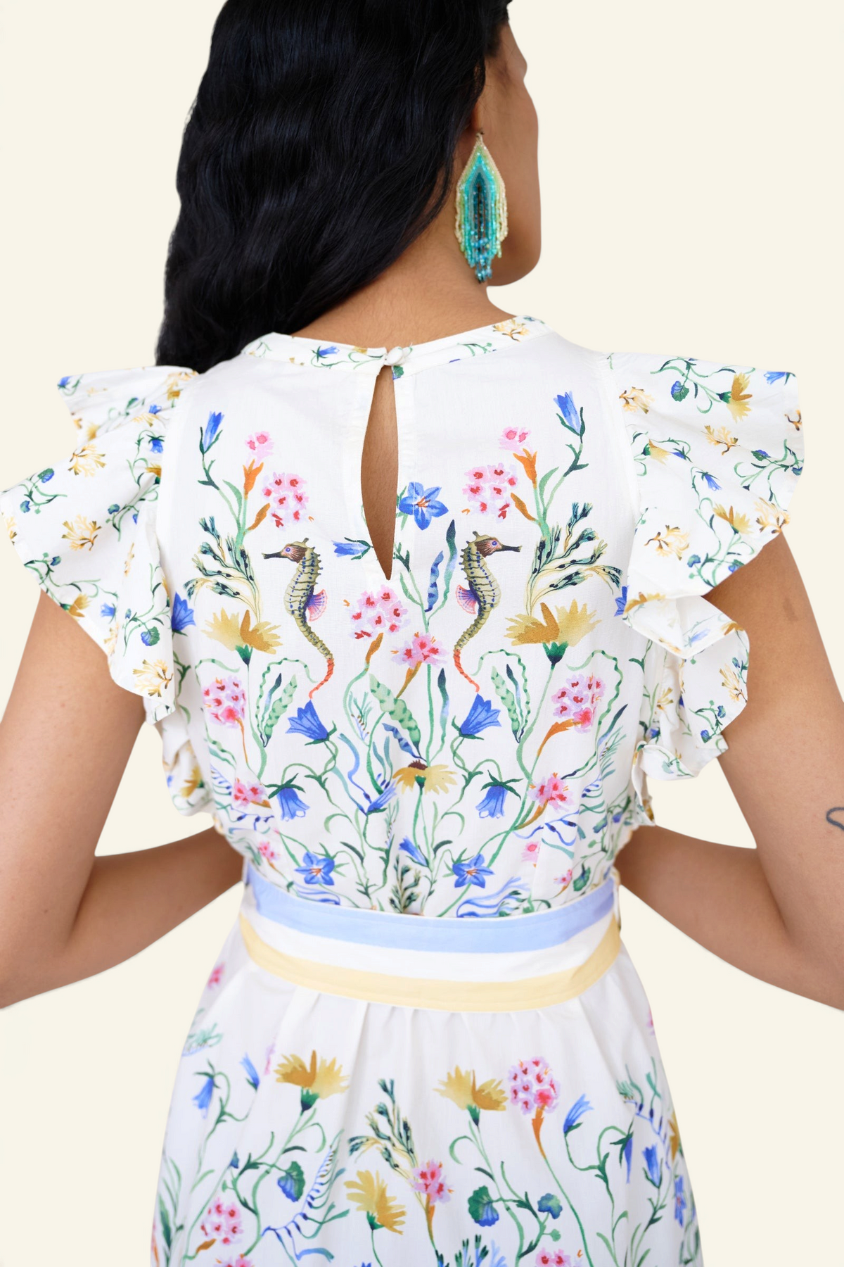 Back view close up of a woman standing. She is wearing the Bulbul dress in Tidepool print. It features ruffled shoulders, removable belt, keyhole back, side-seam pockets.
