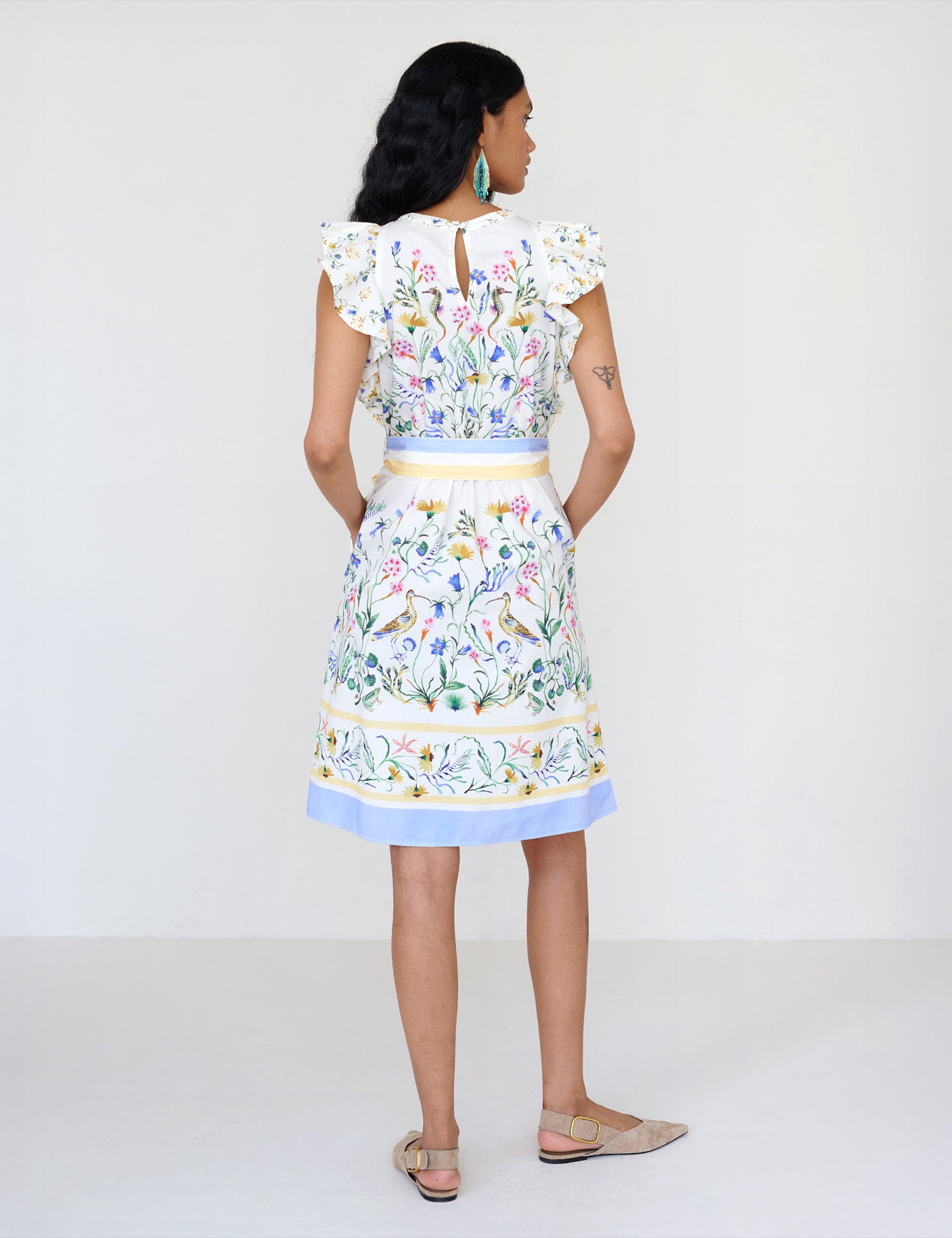 Back view of a woman standing. She is wearing the Bulbul dress in Tidepool print. It features ruffled shoulders, a soft hourglass silhouette, a removable self-belt with blue and yellow stripe, and side-seam pockets.