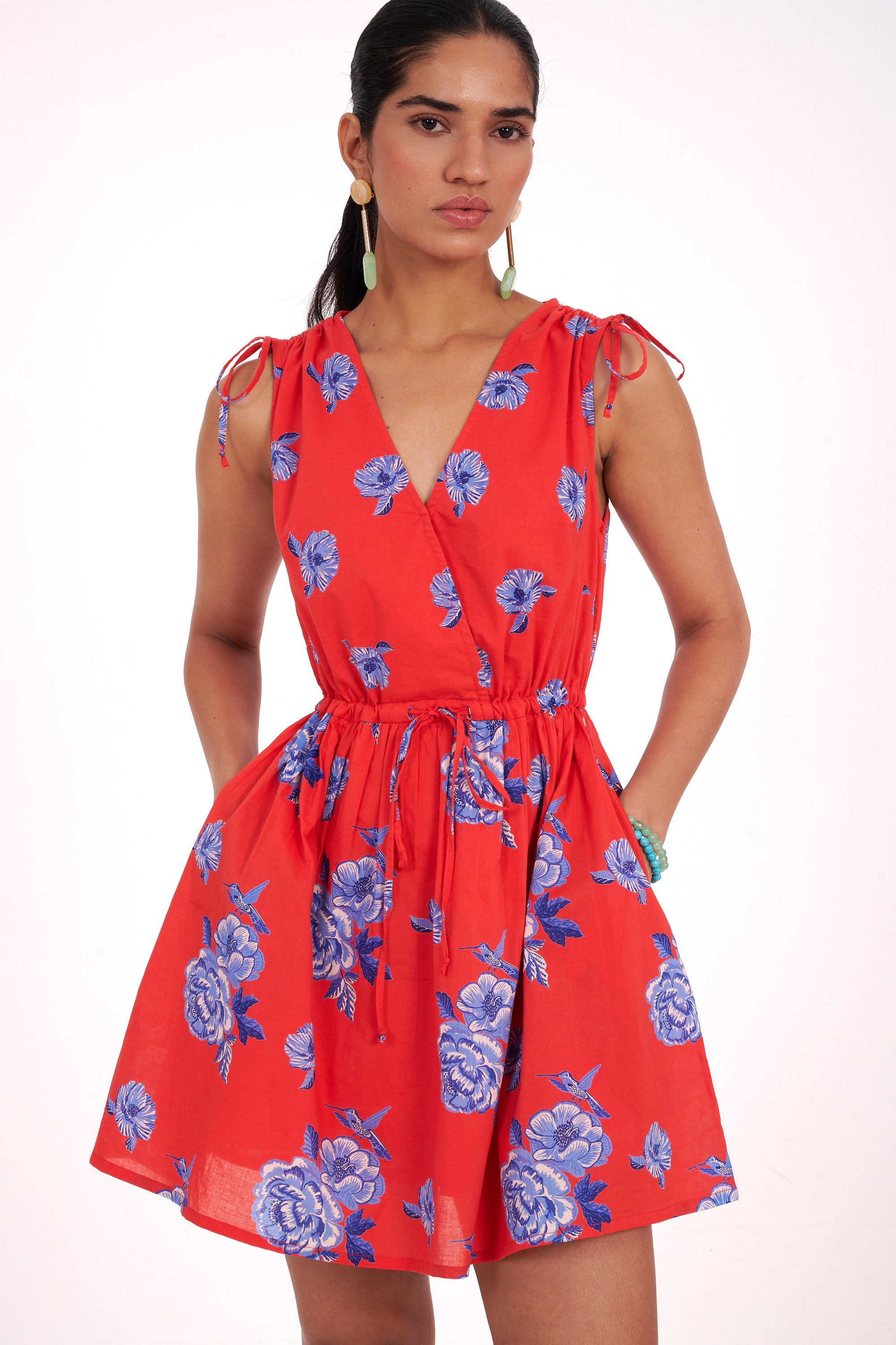 Front view close up of woman standing. She is wearing the Becca dress in the Eliza Rose print, featuring a mini-dress silhouette, adjustable waist, shoulder ties, and floral motifs with blue roses and hummingbirds.
