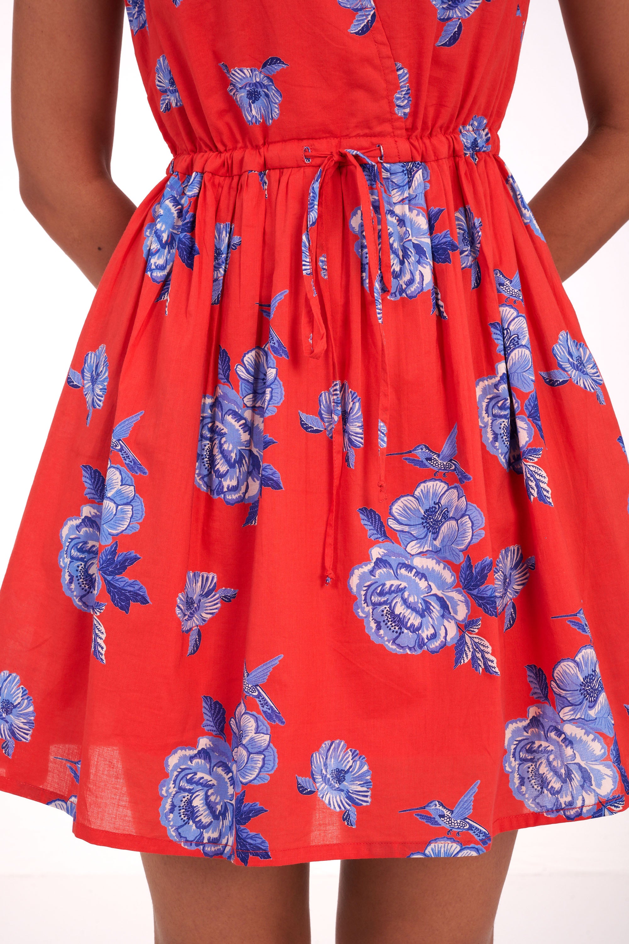 Front view close up of woman standing. She is wearing the Becca dress in the Eliza Rose print, featuring a mini-dress silhouette, adjustable waist, shoulder ties, and floral motifs with blue roses and hummingbirds.