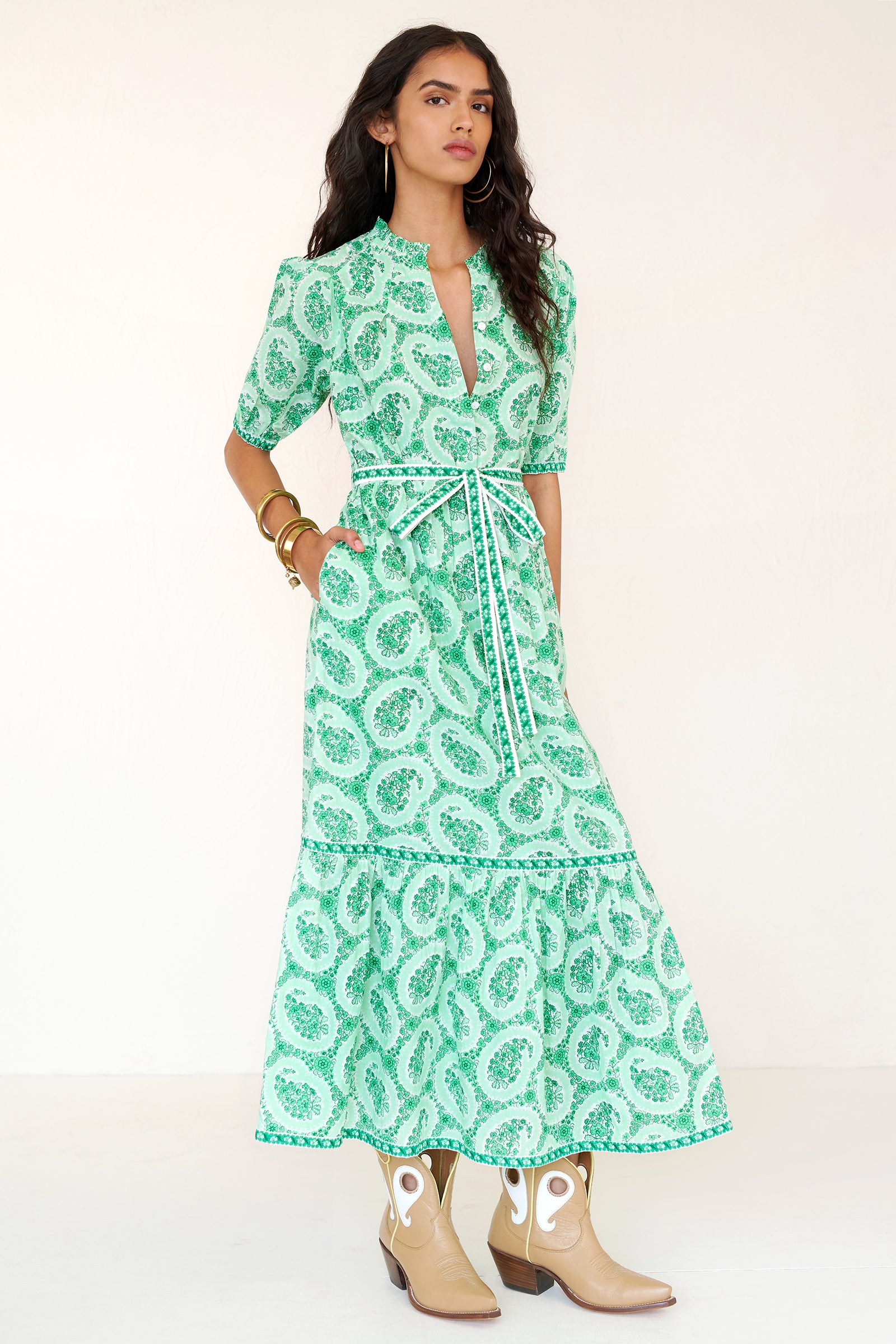 Front side view of a woman standing wearing the Betty dress in Paisley Copse print. It features gathered skirt, raglan sleeves, contrast trim, fabric tie belt, and side-seam pockets in breezy cotton voile.