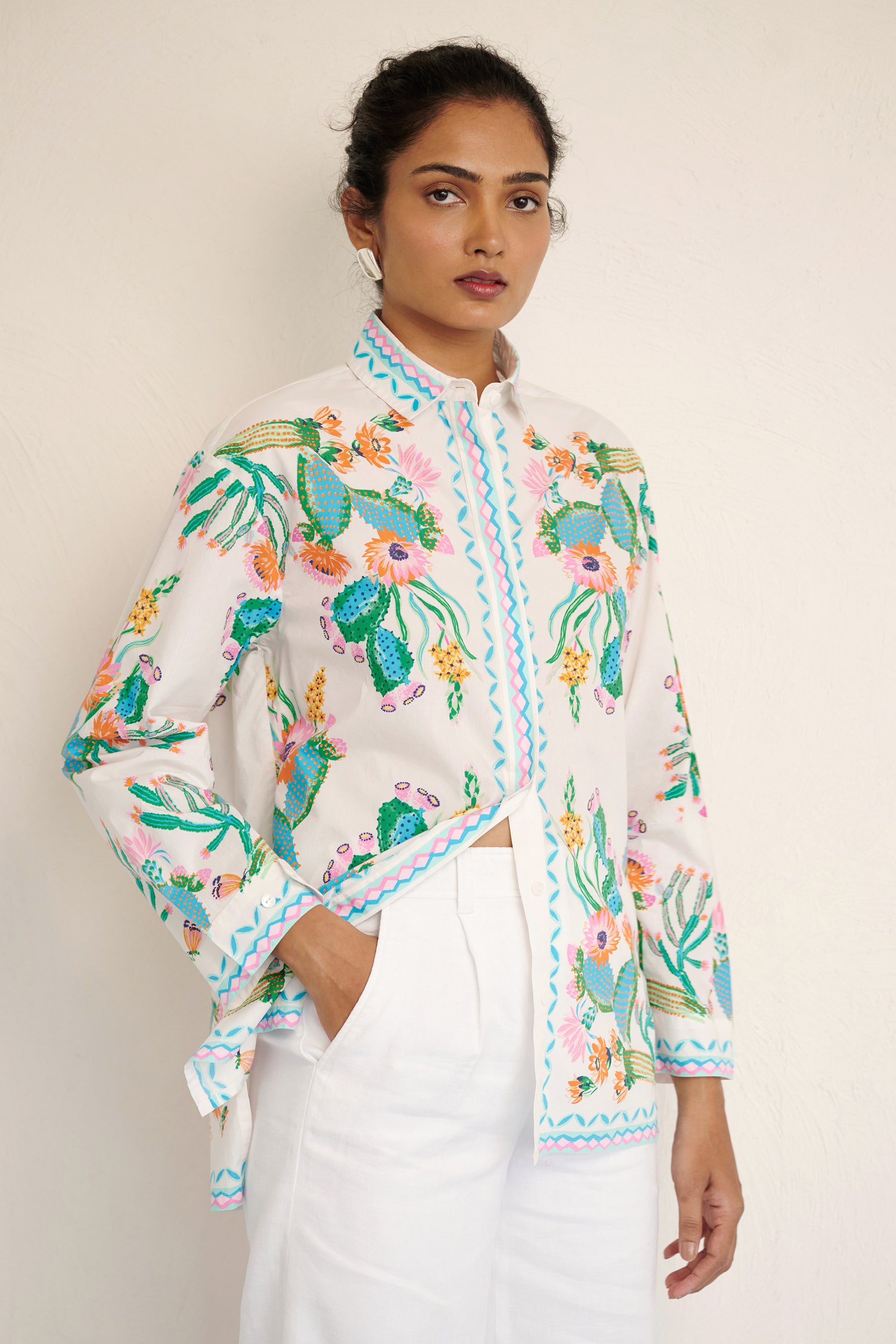 Front side view of woman wearing the Charlotte shirt in Calypso Cacti print, featuring a button-up front, high-low hem, and a back box pleat, printed with cactus blossoms and geometric borders.