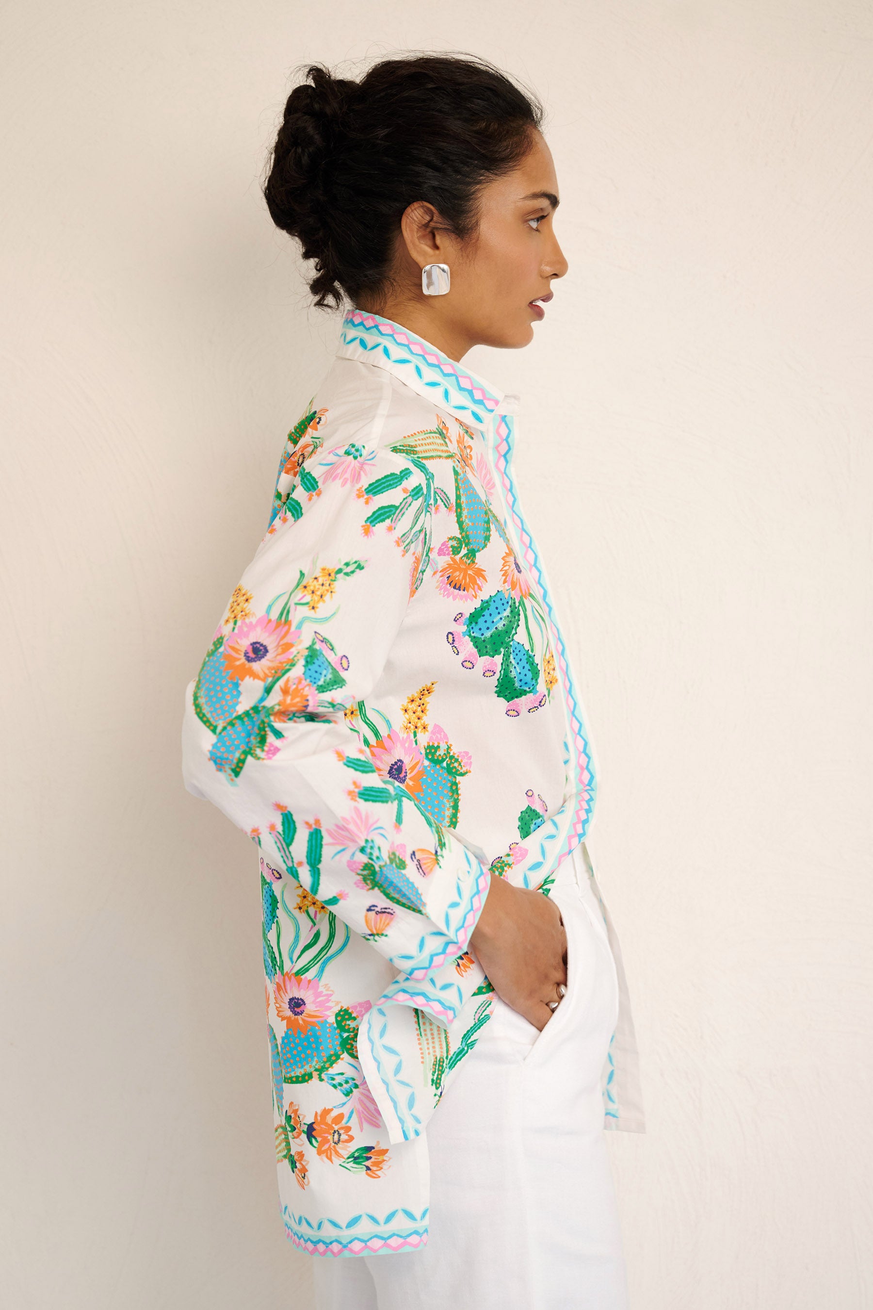 Side view of woman wearing the Charlotte shirt in Calypso Cacti print, featuring a button-up front, high-low hem, and a back box pleat, printed with cactus blossoms and geometric borders.