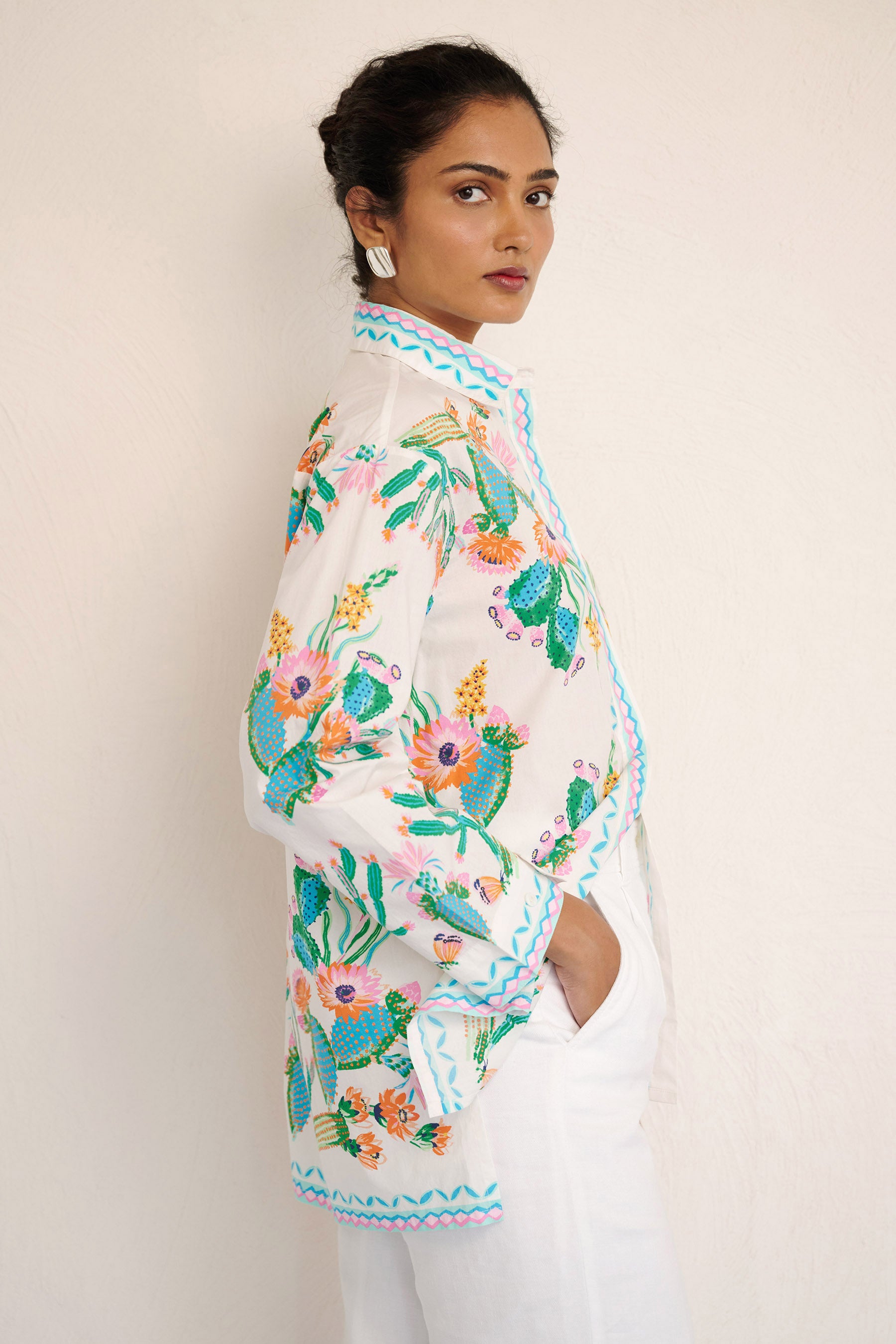 Side view of woman wearing the Charlotte shirt in Calypso Cacti print, featuring a button-up front, high-low hem, and a back box pleat, printed with cactus blossoms and geometric borders.