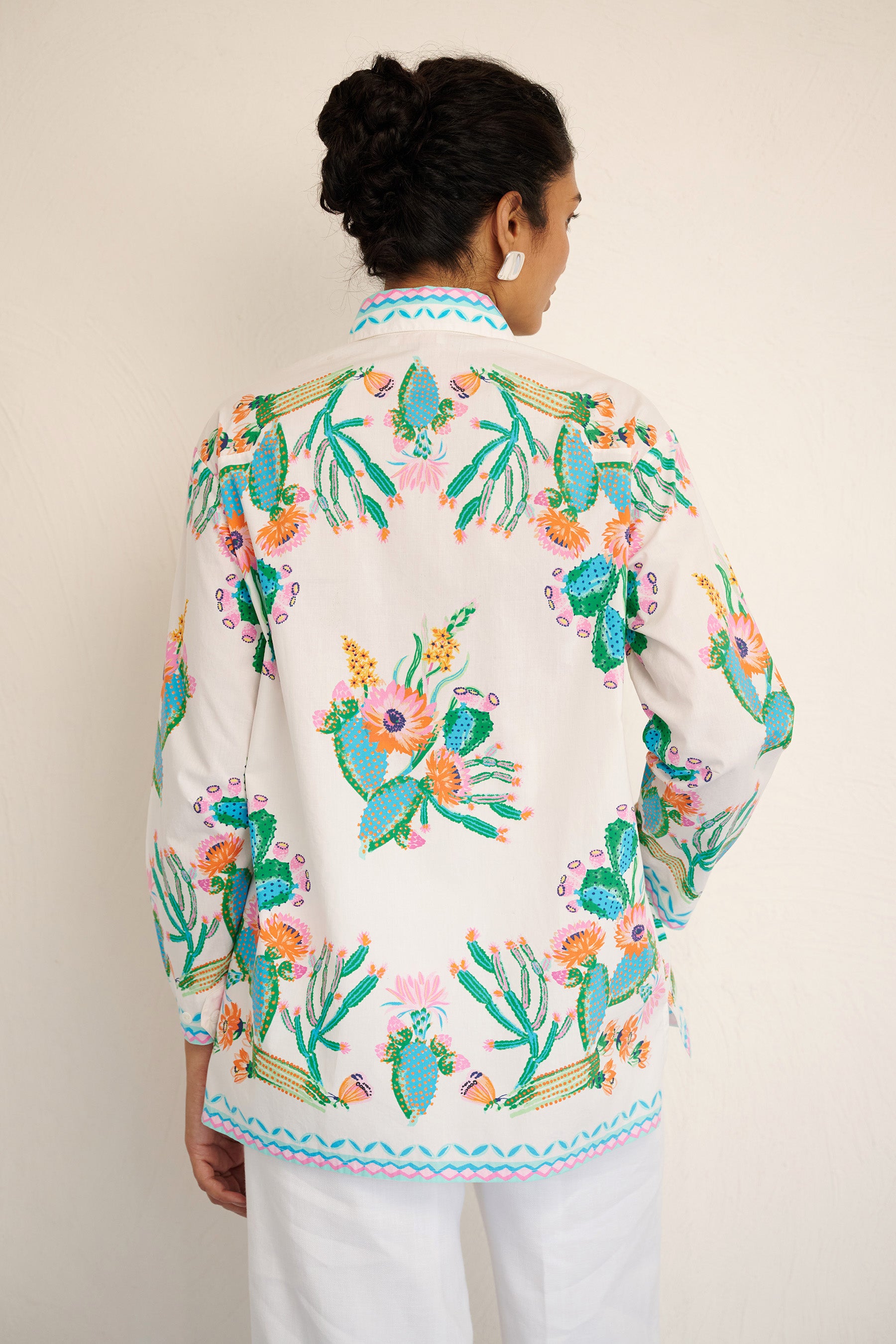 Back view of woman wearing the Charlotte shirt in Calypso Cacti print, featuring a button-up front, high-low hem, and a back box pleat, printed with cactus blossoms and geometric borders.
