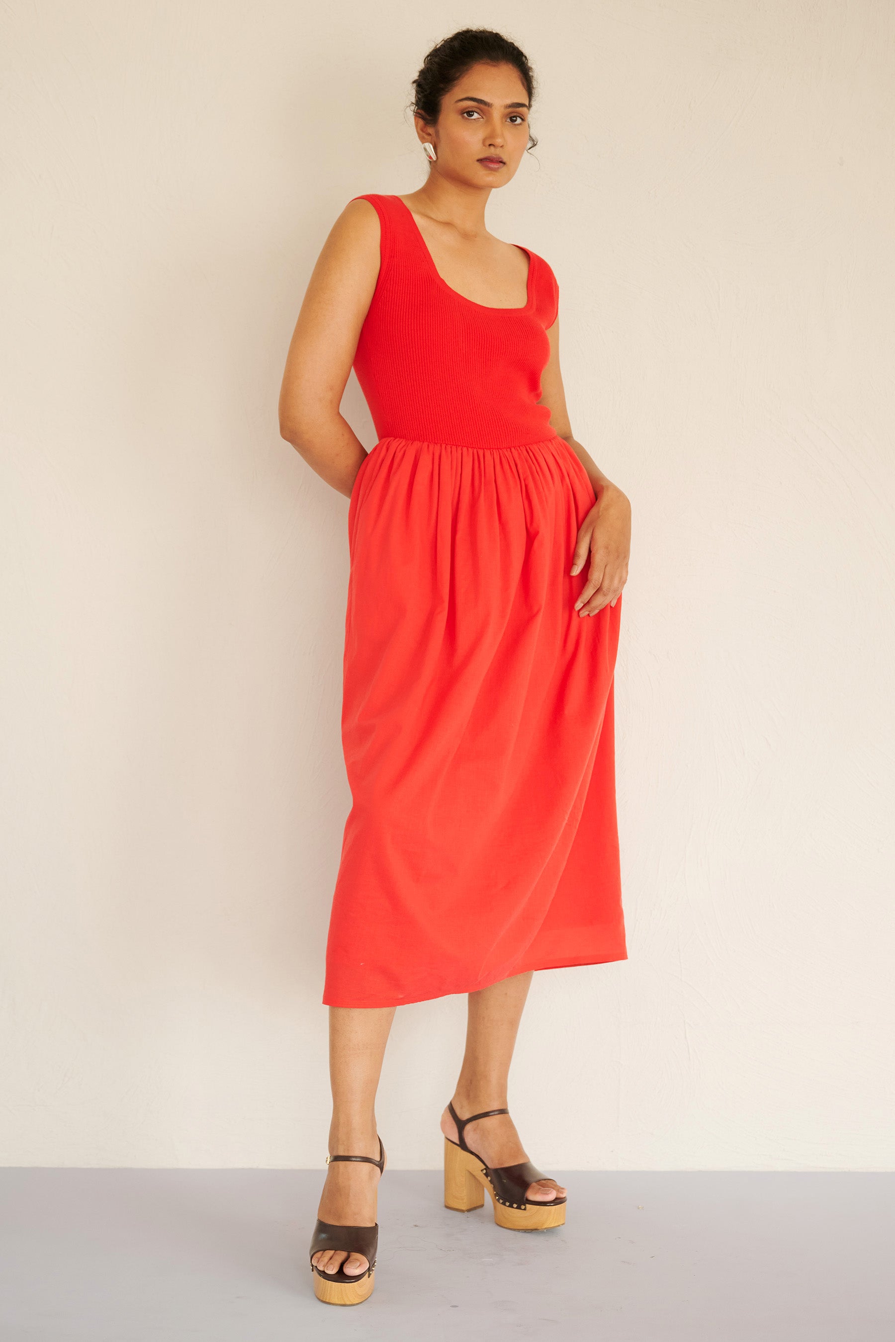 Front view of woman standing. She is wearing the Chloe dress in poppy red, featuring a ribbed square-neck knit bodice and a tonal cotton voile gathered skirt, styled with platform heels.