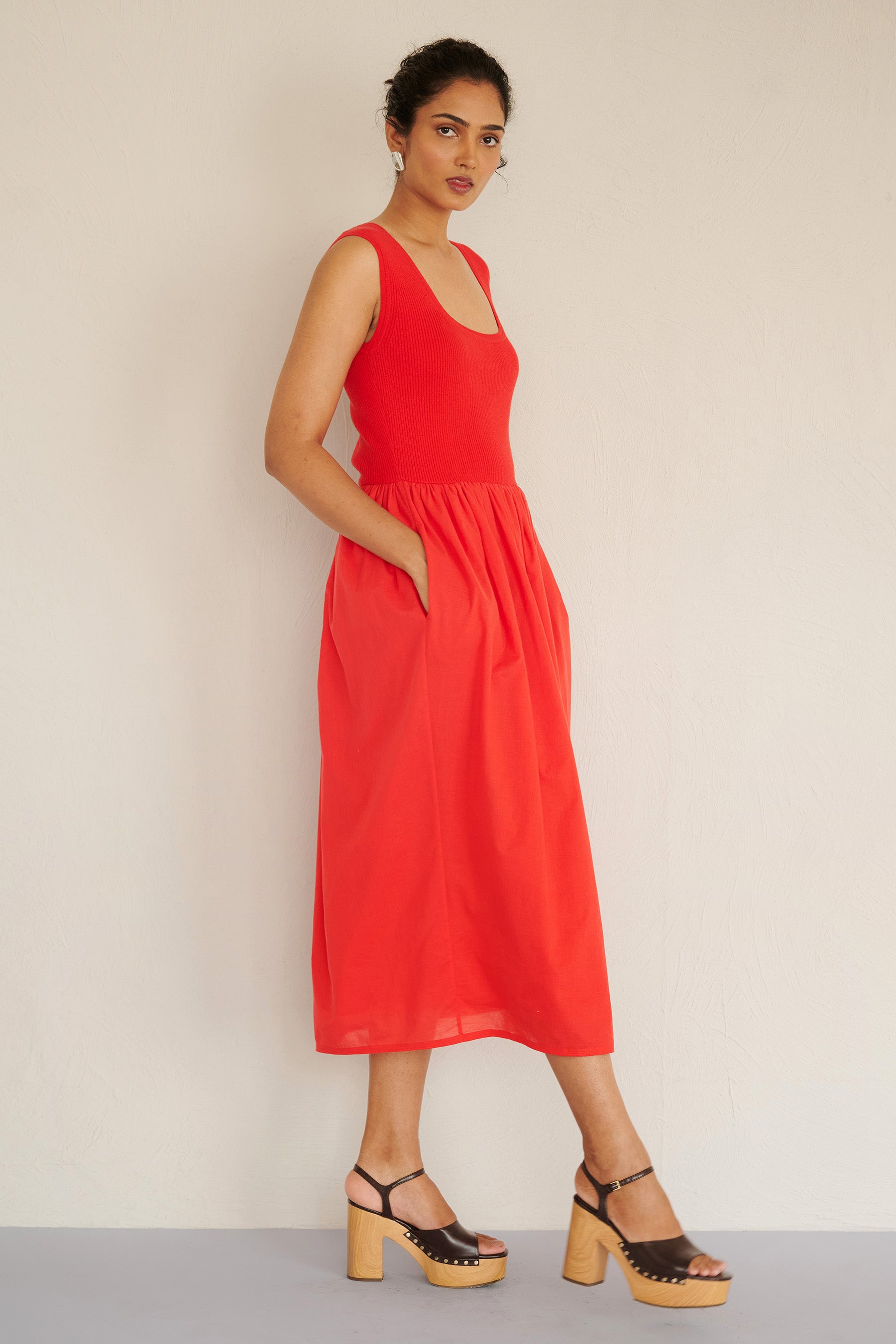 Side view of woman standing. She is wearing the Chloe dress in poppy red, featuring a ribbed square-neck knit bodice and a tonal cotton voile gathered skirt, styled with platform heels.