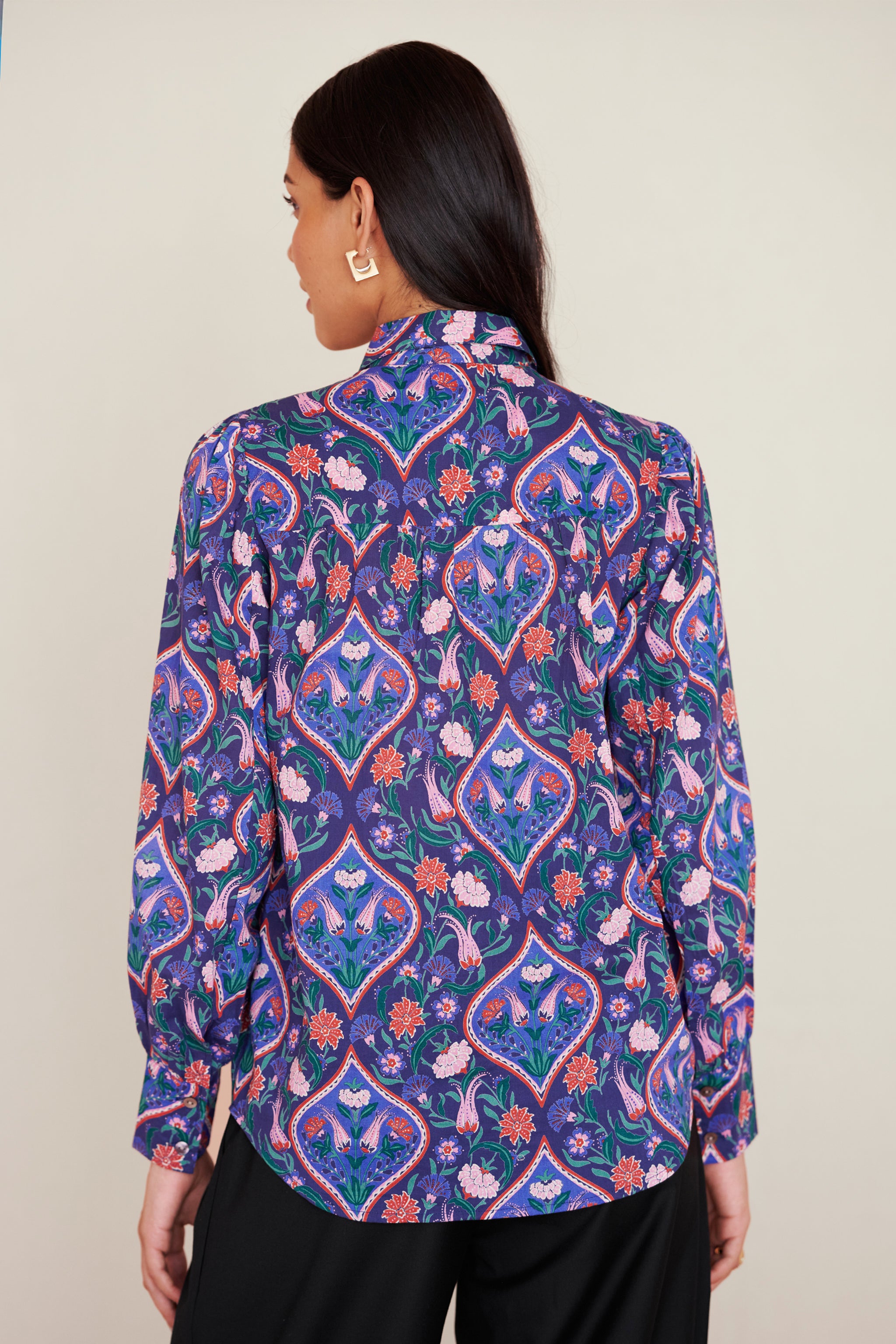 Back view of woman wearing a relaxed-fit button-down shirt with floral tile print, straight collar, and full sleeves with buttoned cuffs.