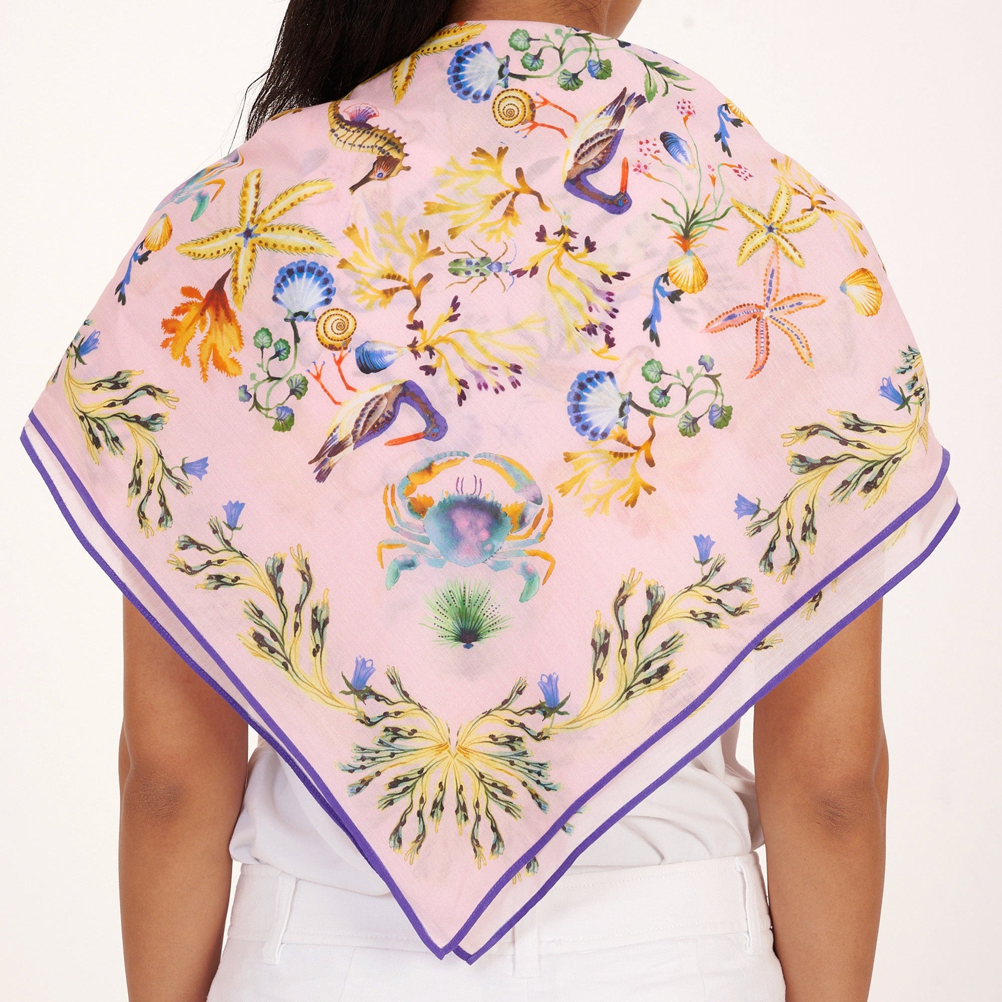 Back view of woman standing, styled with the Carolina scarf draped over her shoulder in the Tidepool print featuring starfish, seahorse, and shell motifs, paired with white pants.