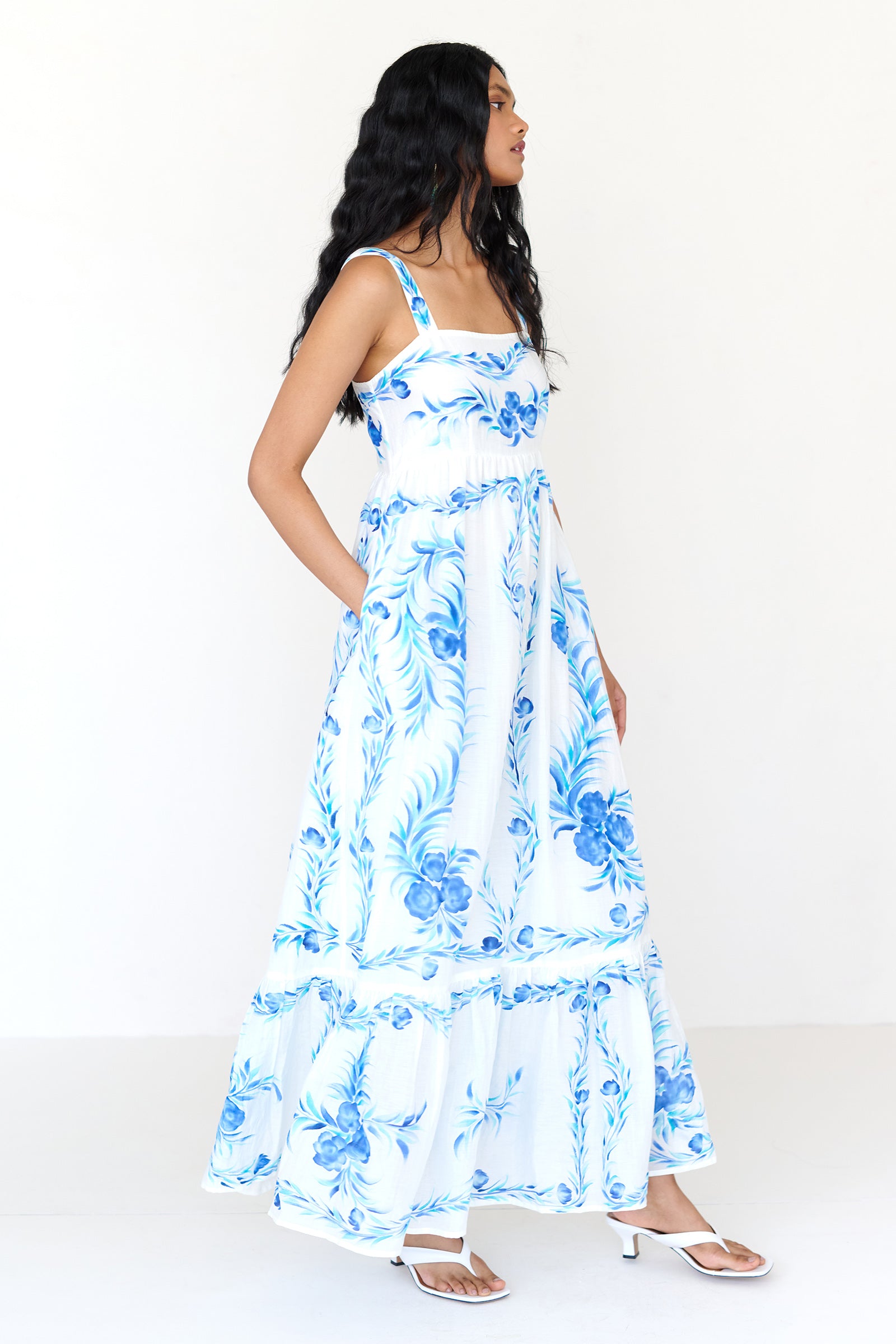 Side view of a woman standing wearing the Daniella dress, featuring hand-painted floral vignettes, ankle-grazing hem, straight neckline and side-seam pockets.