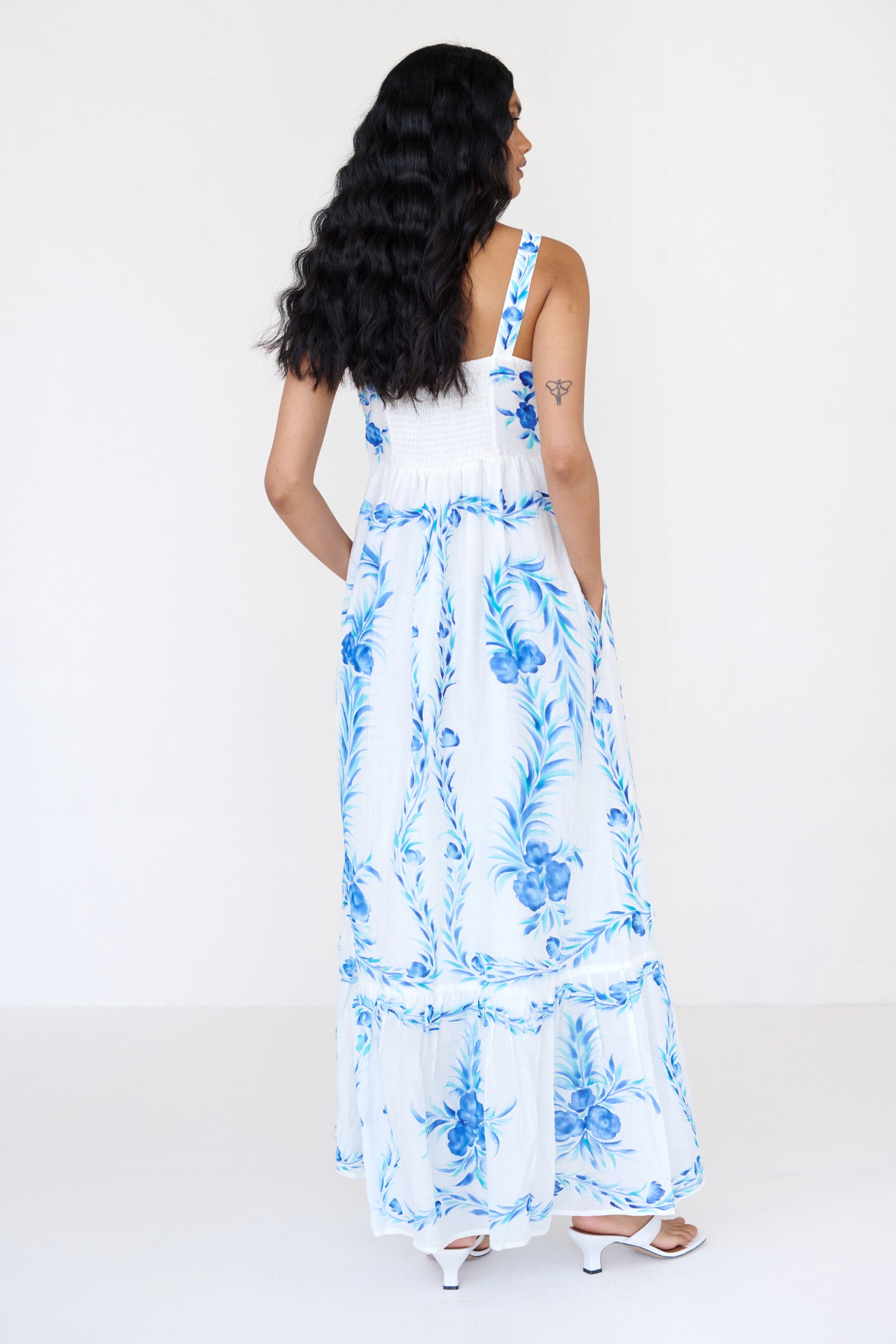 Back view of a woman standing wearing the Daniella dress, featuring hand-painted floral vignettes, ankle-grazing hem, straight neckline and side-seam pockets.