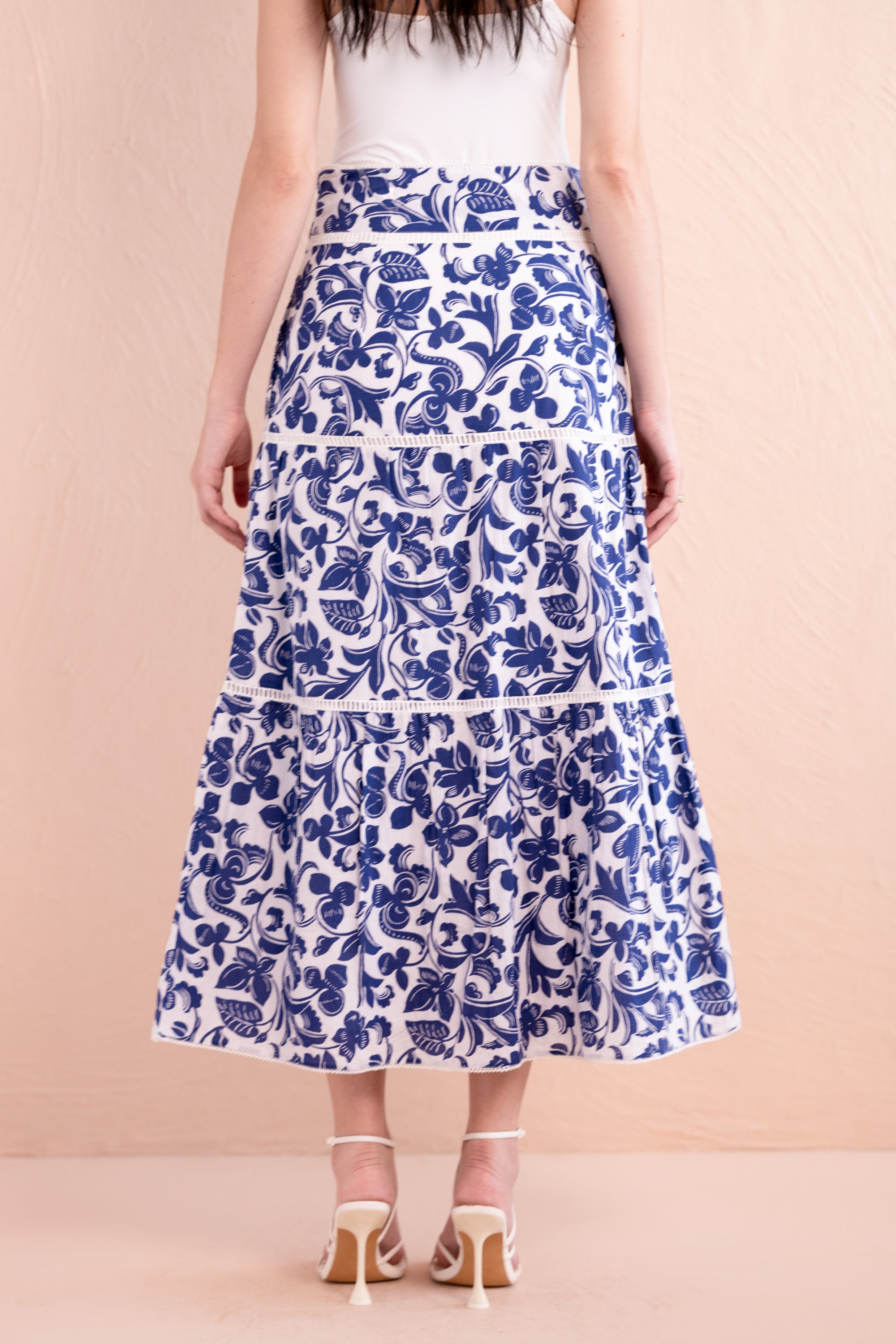 Back view of a woman wearing a white and cobalt blue floral printed tiered midi skirt with pockets, styled with a white top and strappy white sandals.