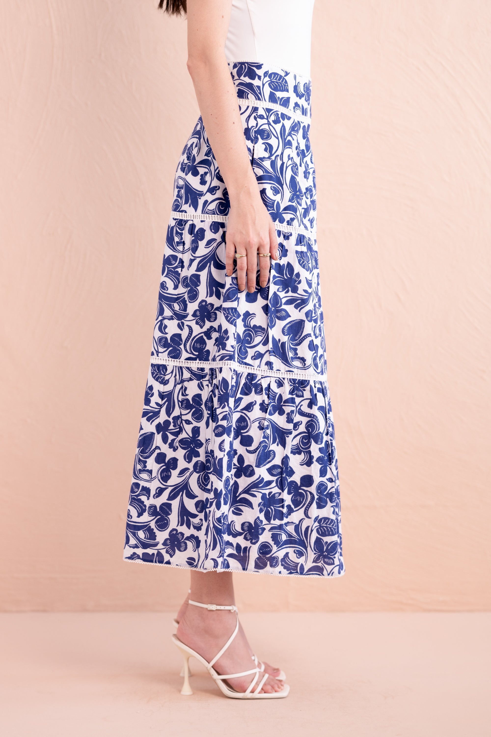 Side view of a woman wearing a white and cobalt blue floral printed tiered midi skirt with pockets, styled with a white top and strappy white sandals.