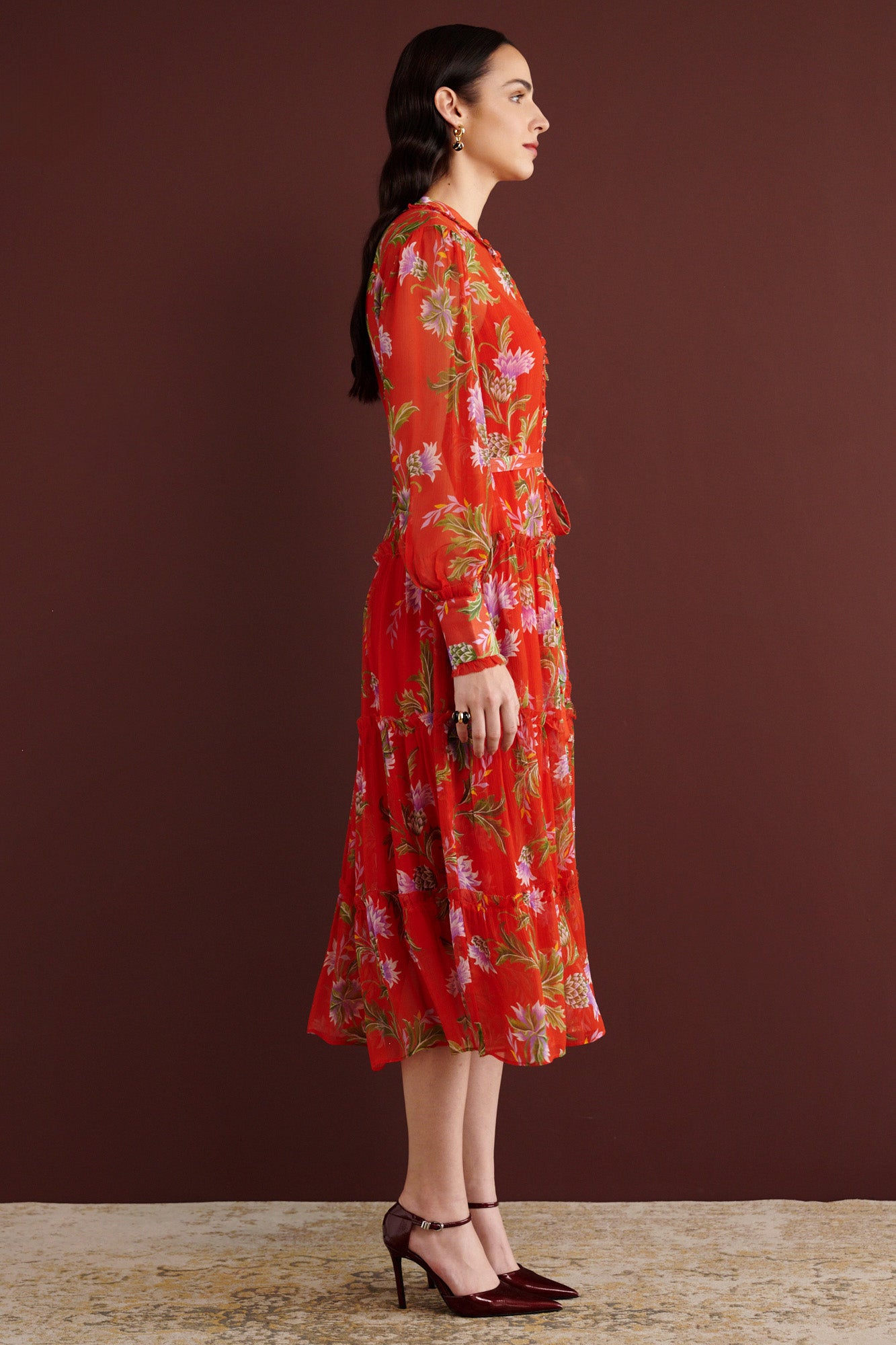 Side view of woman standing. She is wearing the Emira dress in the Stuart Thistle print, a flowing Flame-colored crinkle chiffon dress with sheer sleeves, delicate ruffles, fabric-covered buttons, and a detachable belt, featuring floral motifs inspired by the Scottish Highlands.