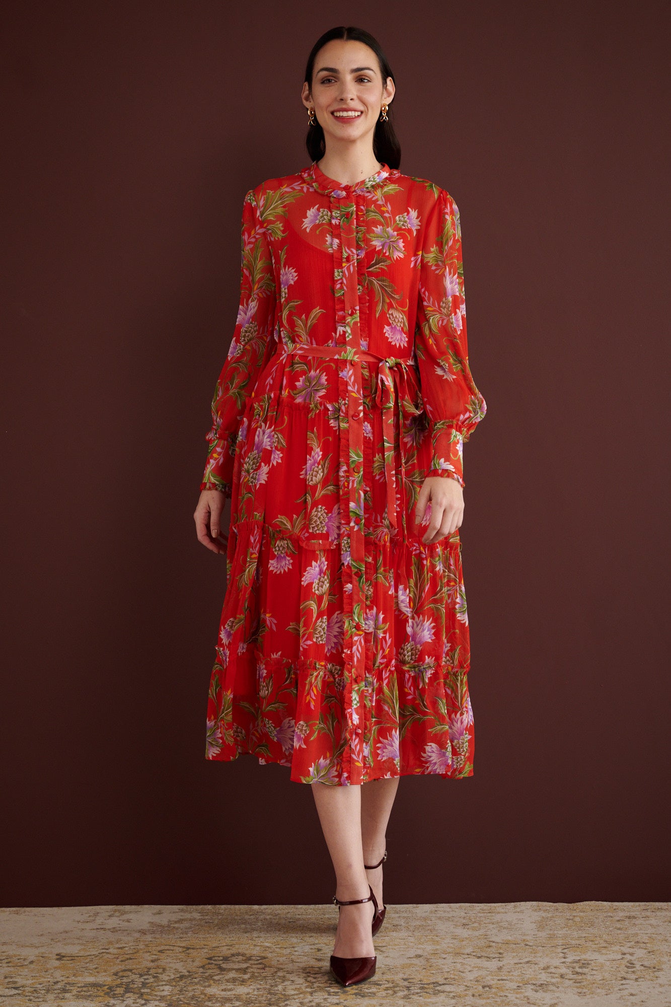 Front view of woman walking. She is wearing the Emira dress in the Stuart Thistle print, a flowing Flame-colored crinkle chiffon dress with sheer sleeves, delicate ruffles, fabric-covered buttons, and a detachable belt, featuring floral motifs inspired by the Scottish Highlands.