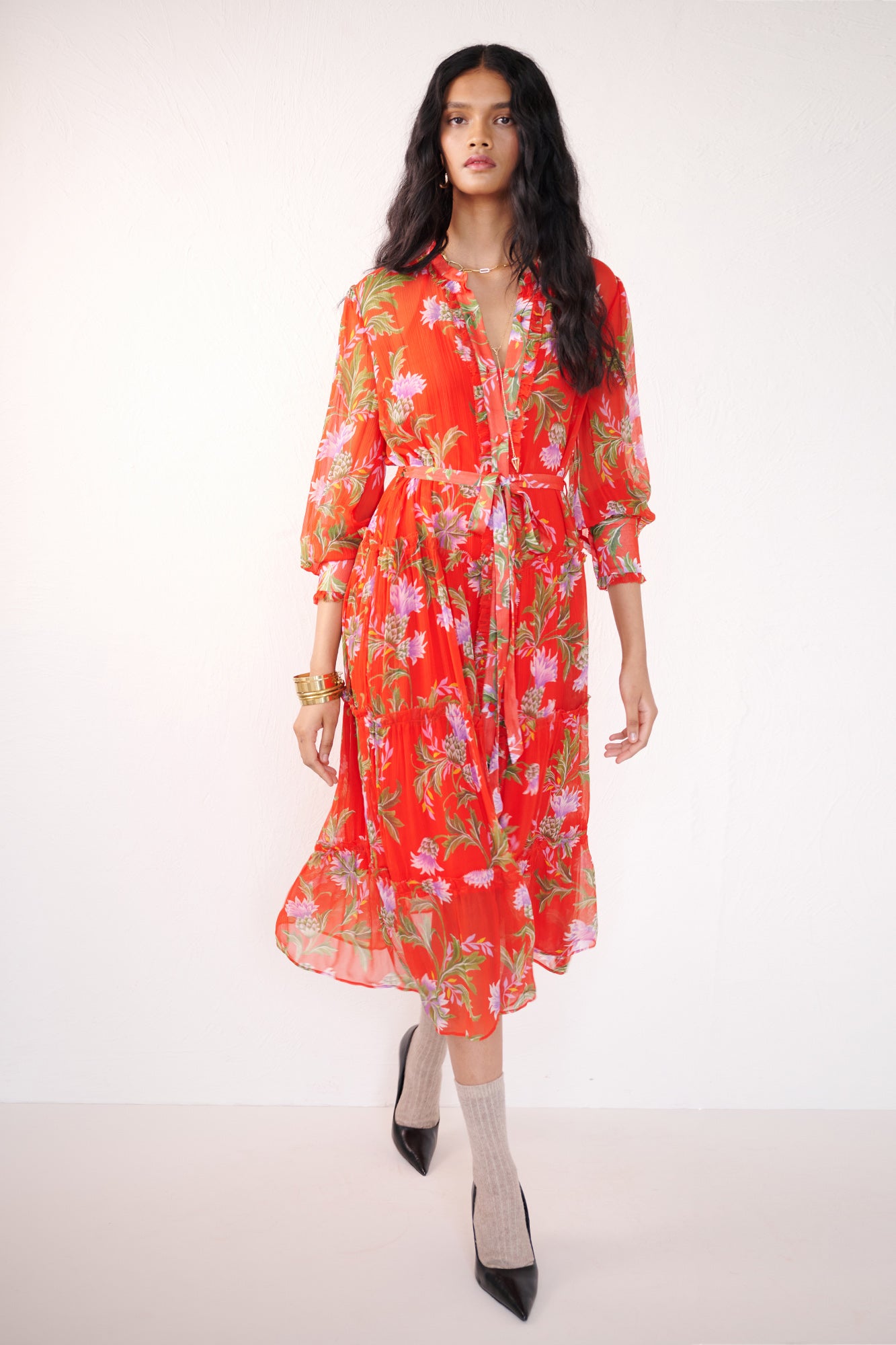 Front view of woman walking. She is wearing the Emira dress in the Stuart Thistle print. It is a flowing Flame-colored crinkle chiffon dress with sheer sleeves, delicate ruffles, fabric-covered buttons, and a detachable belt, featuring floral motifs inspired by the Scottish Highlands.