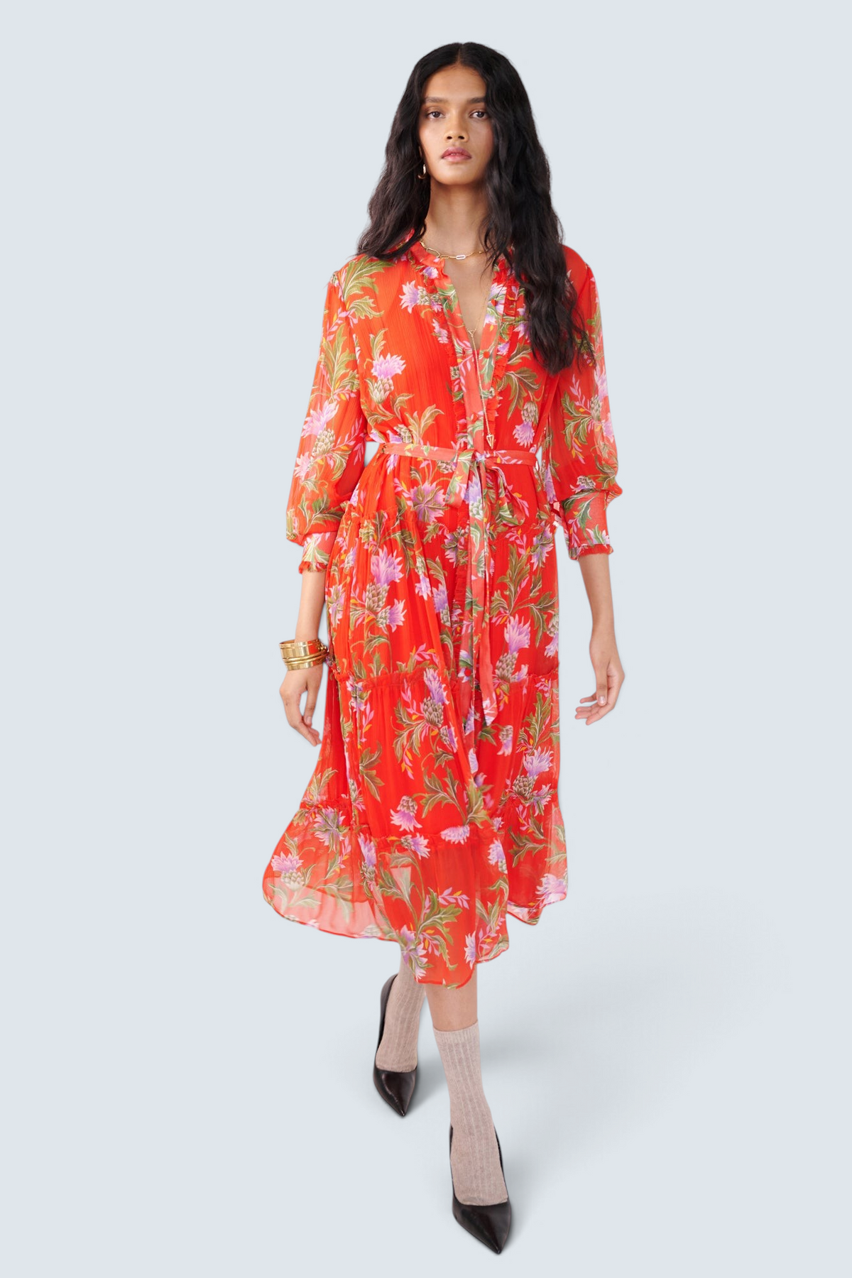 Front view of woman walking. She is wearing the Emira dress in the Stuart Thistle print. It is a flowing Flame-colored crinkle chiffon dress with sheer sleeves, delicate ruffles, fabric-covered buttons, and a detachable belt, featuring floral motifs inspired by the Scottish Highlands.