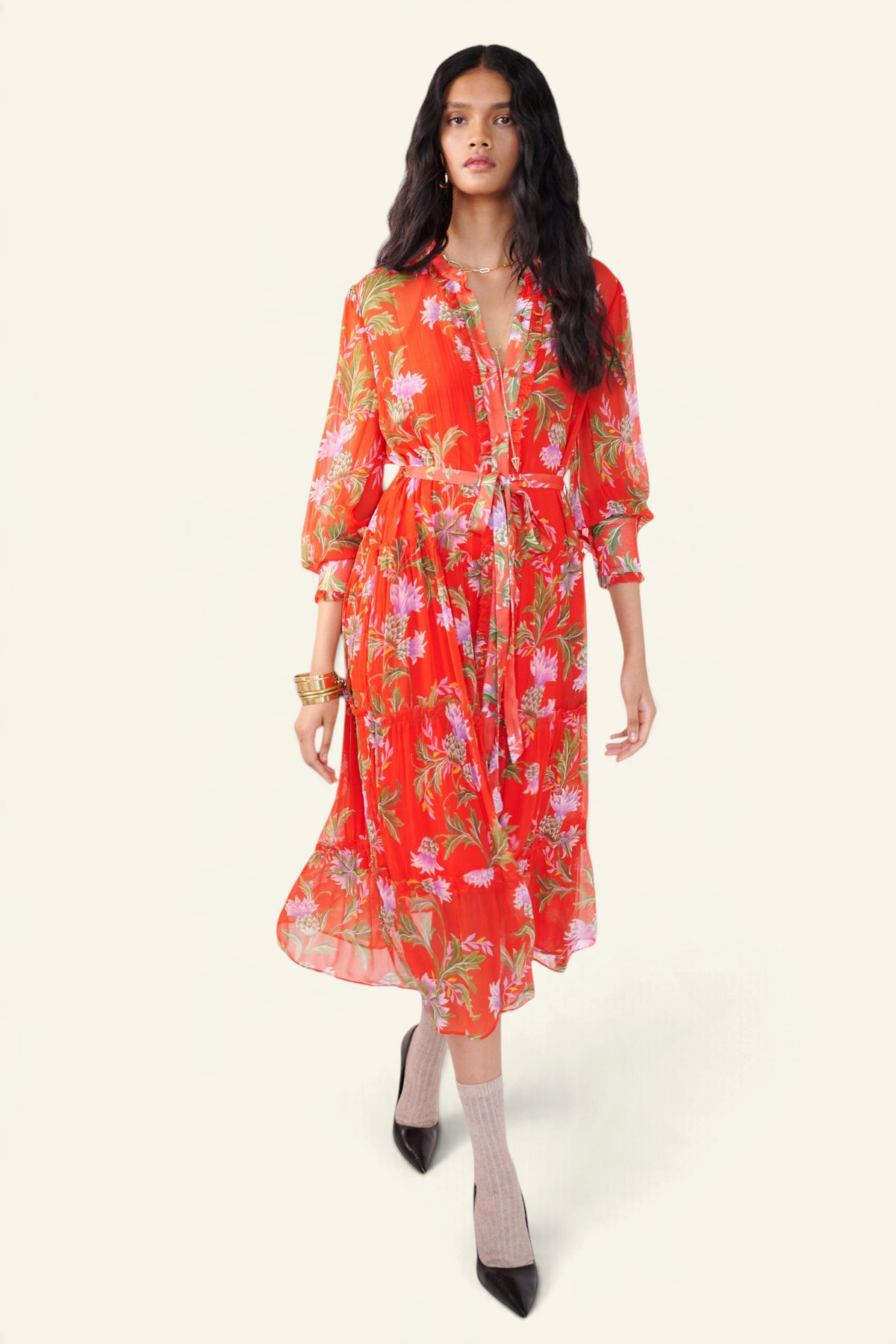 Front view of woman walking. She is wearing the Emira dress in the Stuart Thistle print. It is a flowing Flame-colored crinkle chiffon dress with sheer sleeves, delicate ruffles, fabric-covered buttons, and a detachable belt, featuring floral motifs inspired by the Scottish Highlands.