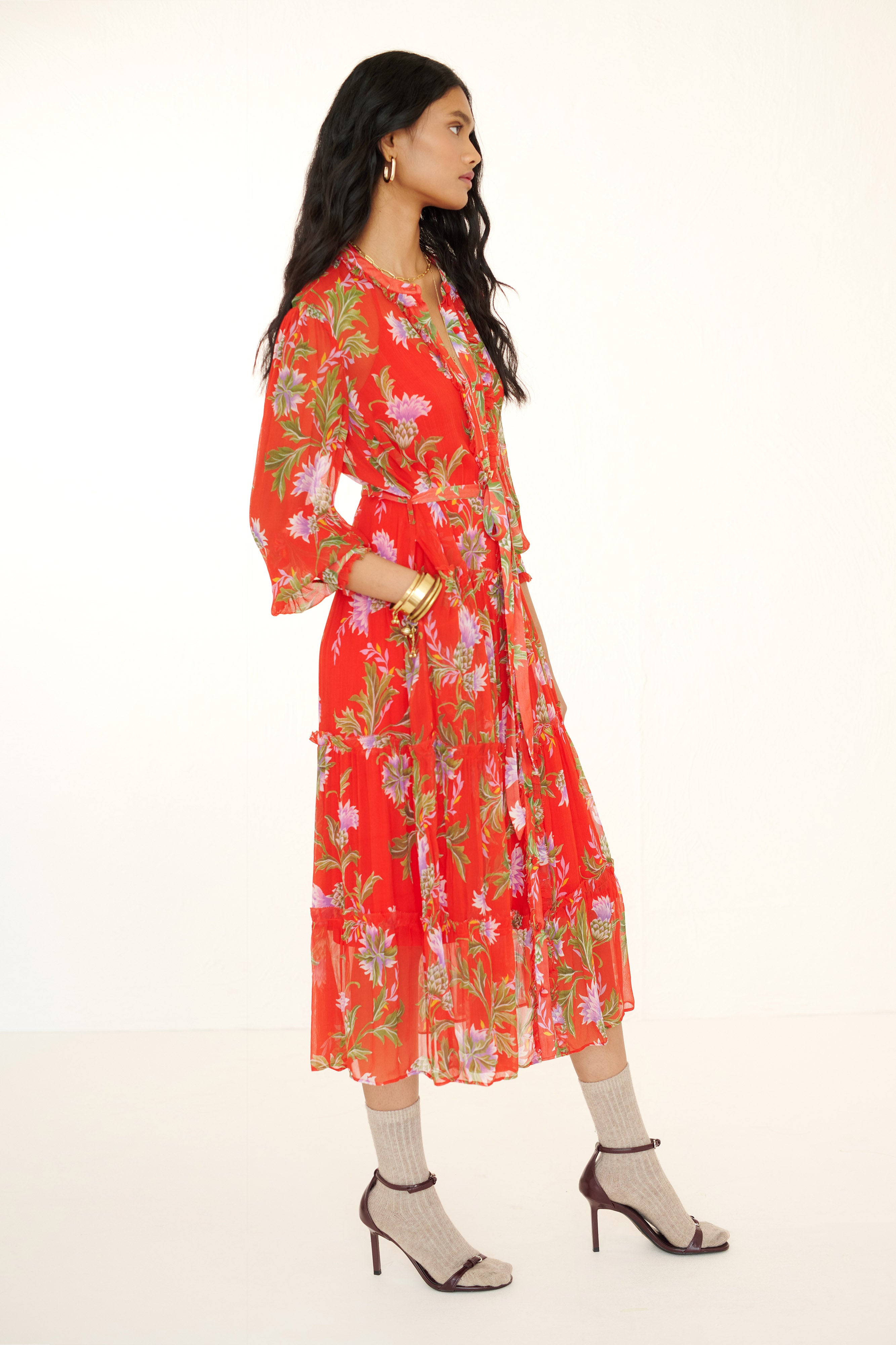 Side view of woman standing with a hand in pocket. She is wearing the Emira dress in the Stuart Thistle print. It is a flowing Flame-colored crinkle chiffon dress with sheer sleeves, delicate ruffles, fabric-covered buttons, and a detachable belt, featuring floral motifs inspired by the Scottish Highlands.